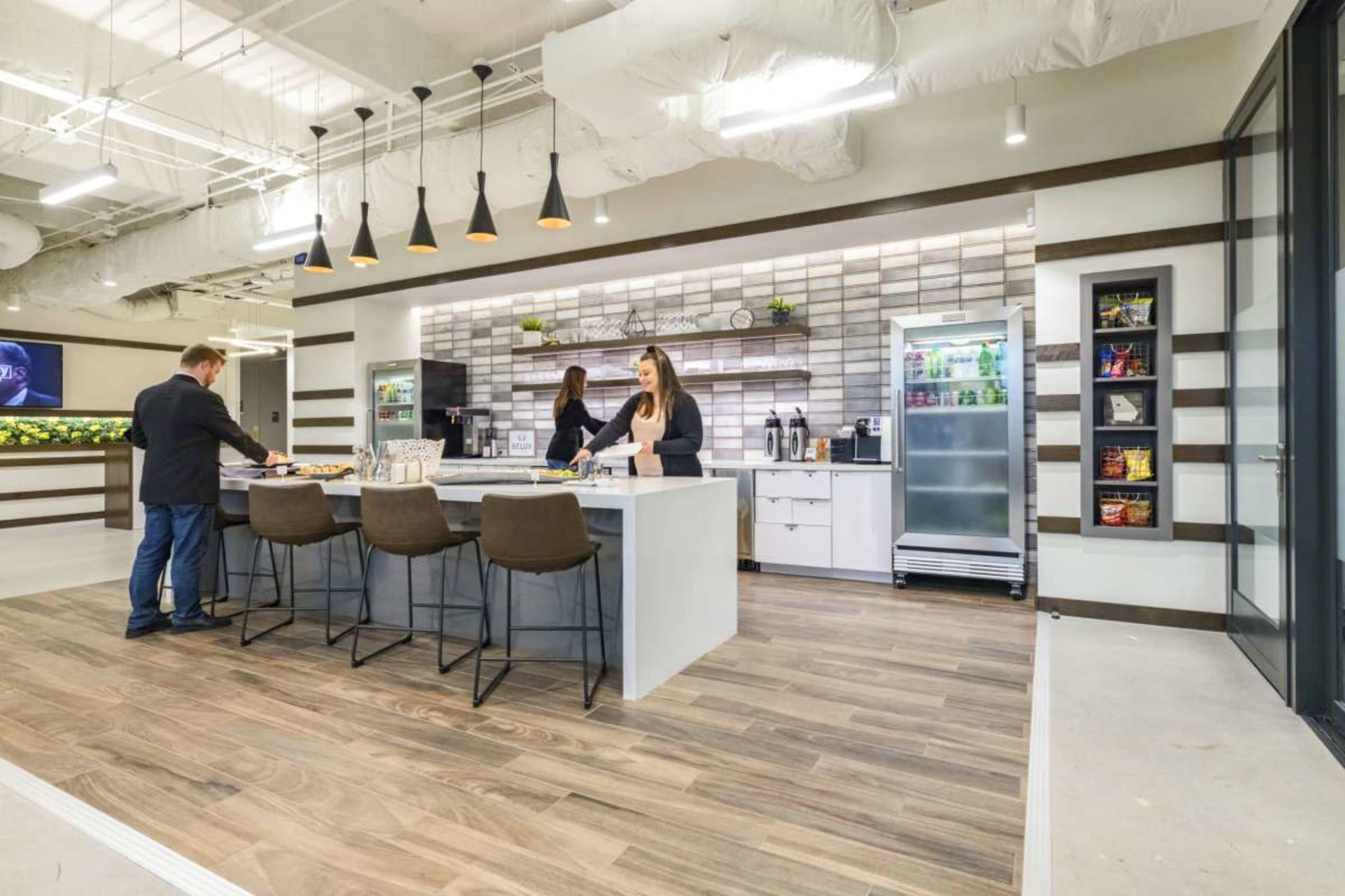The image shows a modern office kitchen area with two people preparing food at a large island, surrounded by sleek cabinets and a refrigerator.
