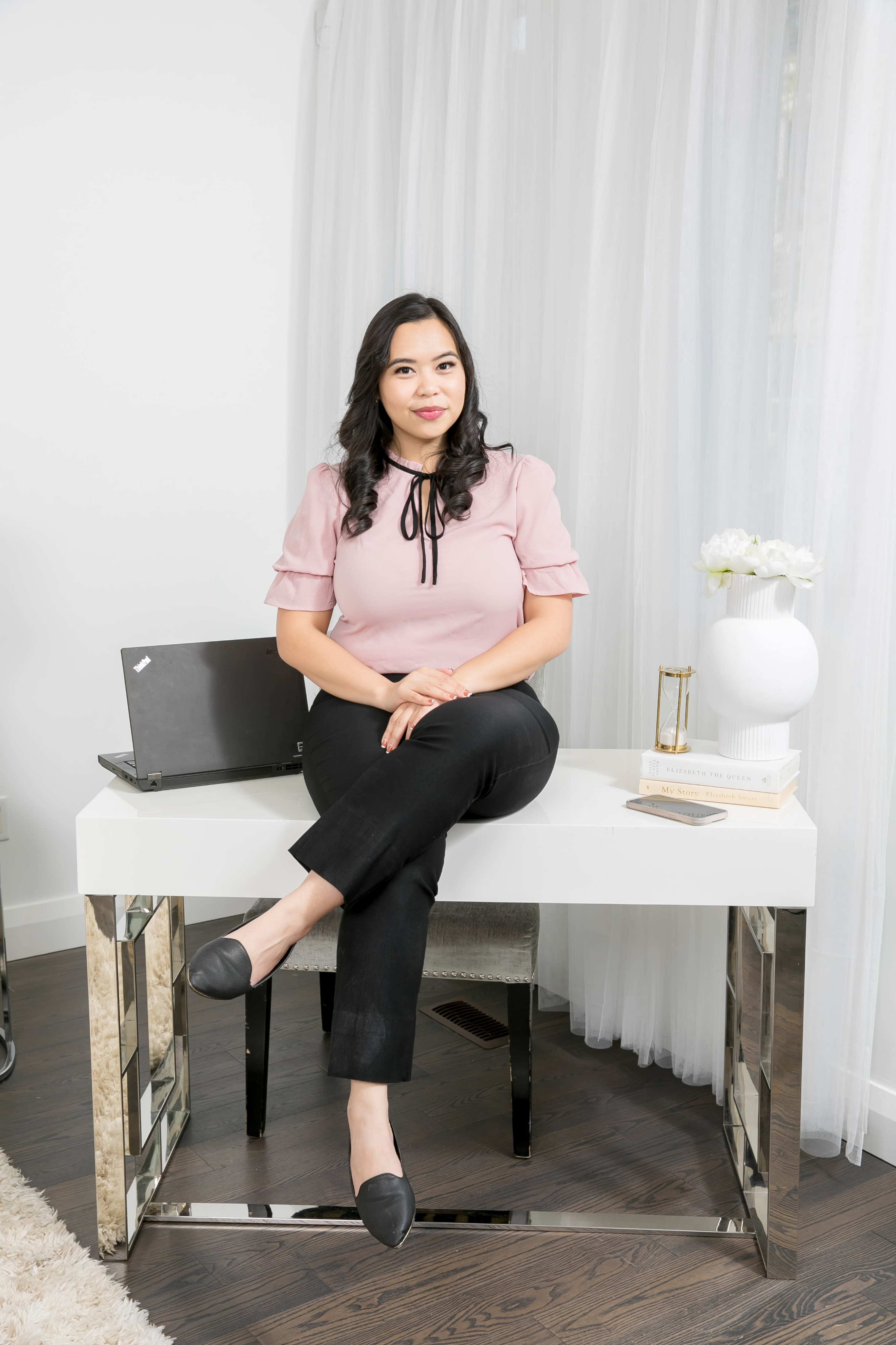 A woman sits at a modern white desk in a bright, airy room, with a laptop and decorative items around her.
