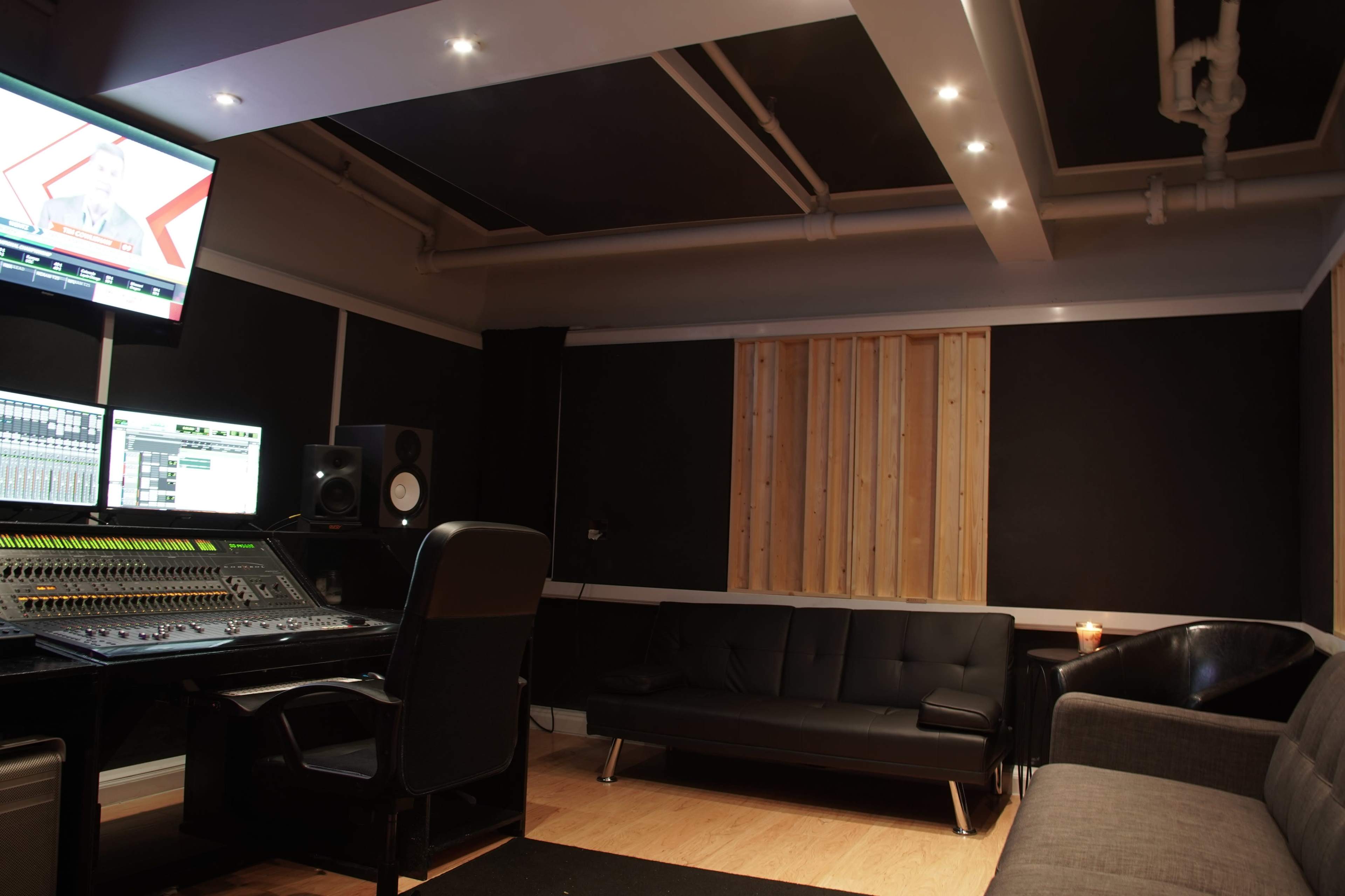 A music recording studio with a mixing console, a television monitor, a black couch, and soundproofed walls.