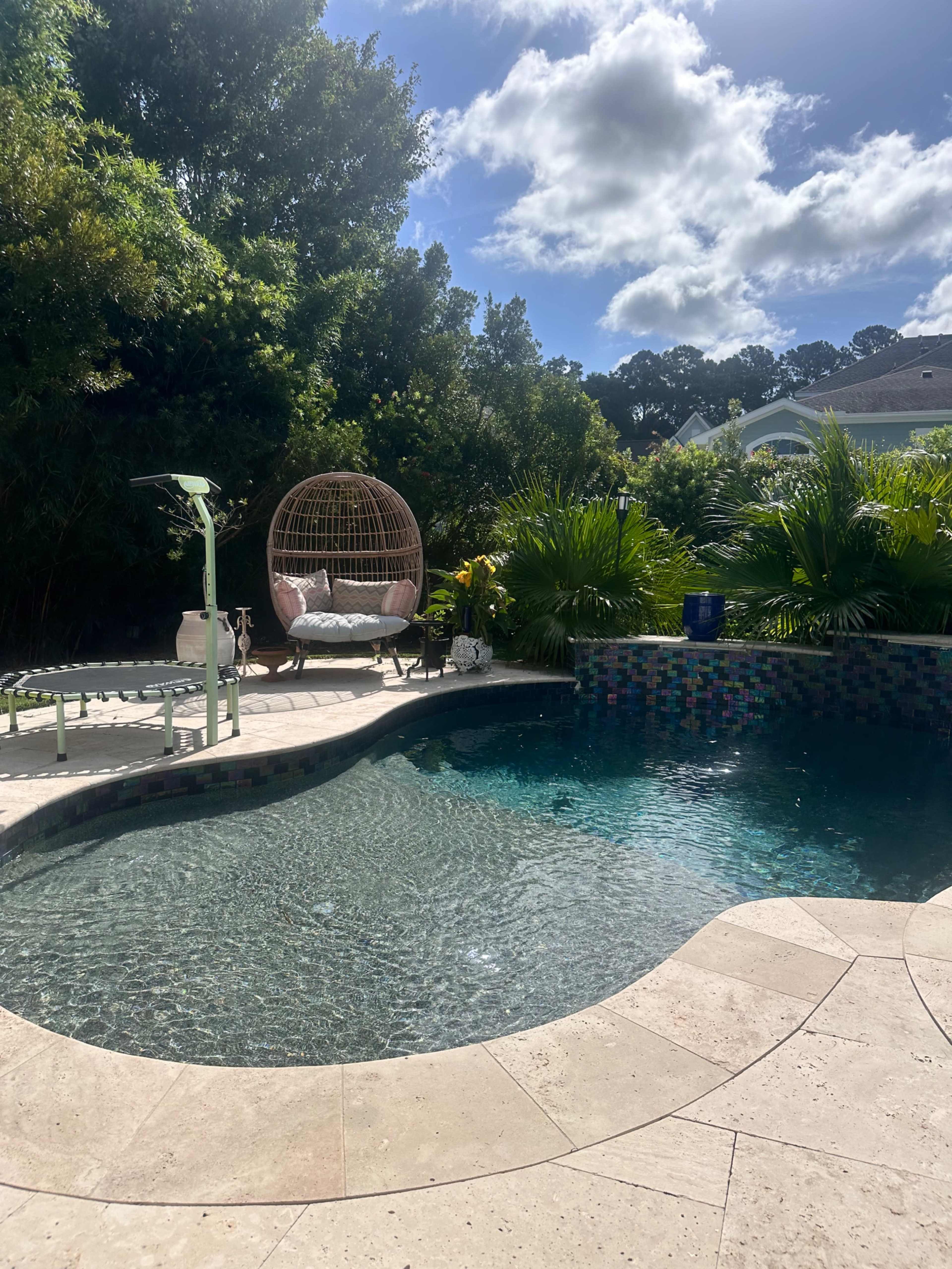 A swimming pool surrounded by lush greenery features a seated hanging chair and a sun lounger.