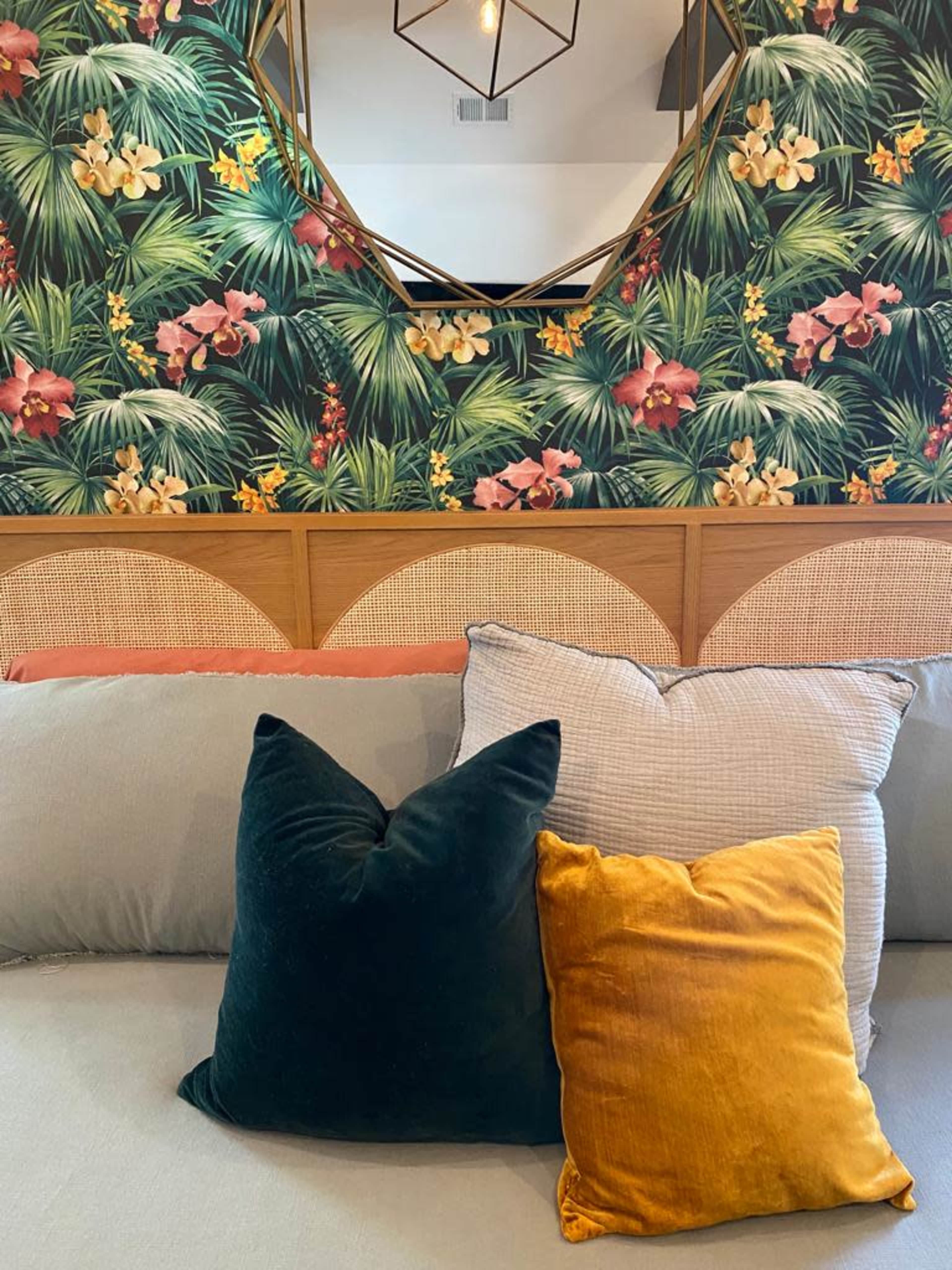 A colorful arrangement of pillows sits on a couch against a backdrop of floral wallpaper and a round mirror.