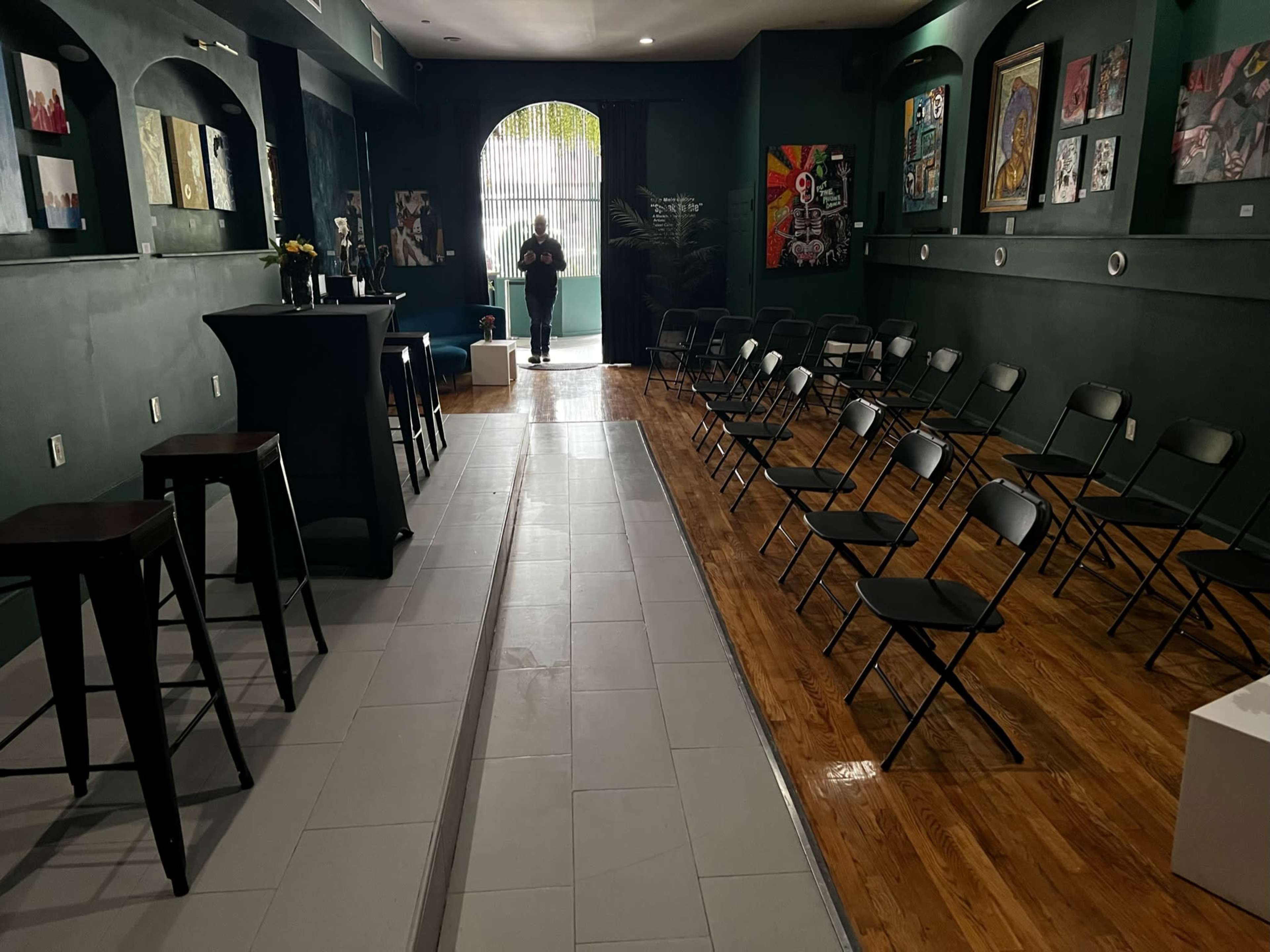 A dimly lit room with an arrangement of black folding chairs facing a bar-like table, framed artwork on the walls, and a person walking towards the entrance.