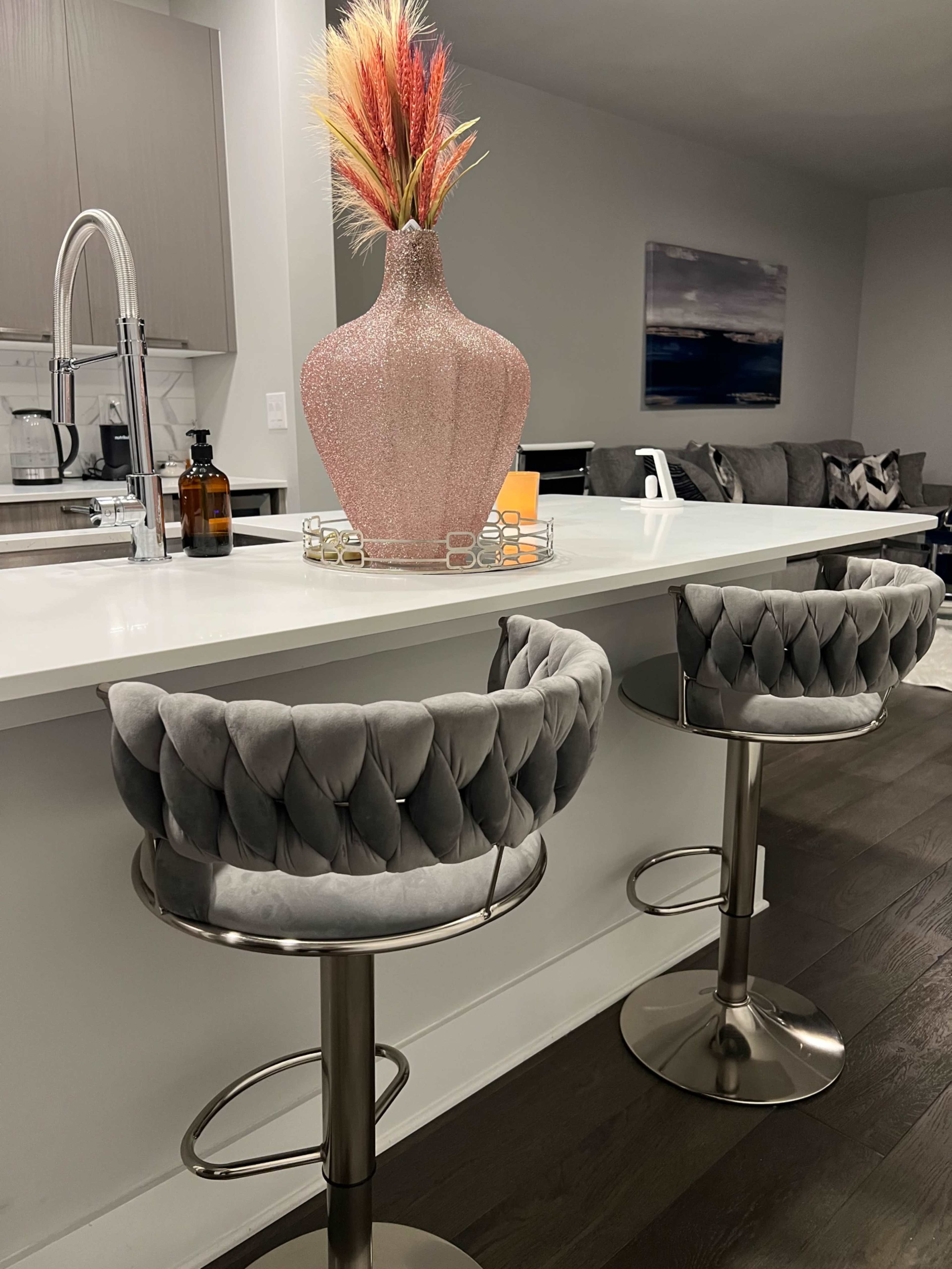 A kitchen bar area features two plush gray stools and a decorative pink vase with colorful dried flowers on a white countertop.