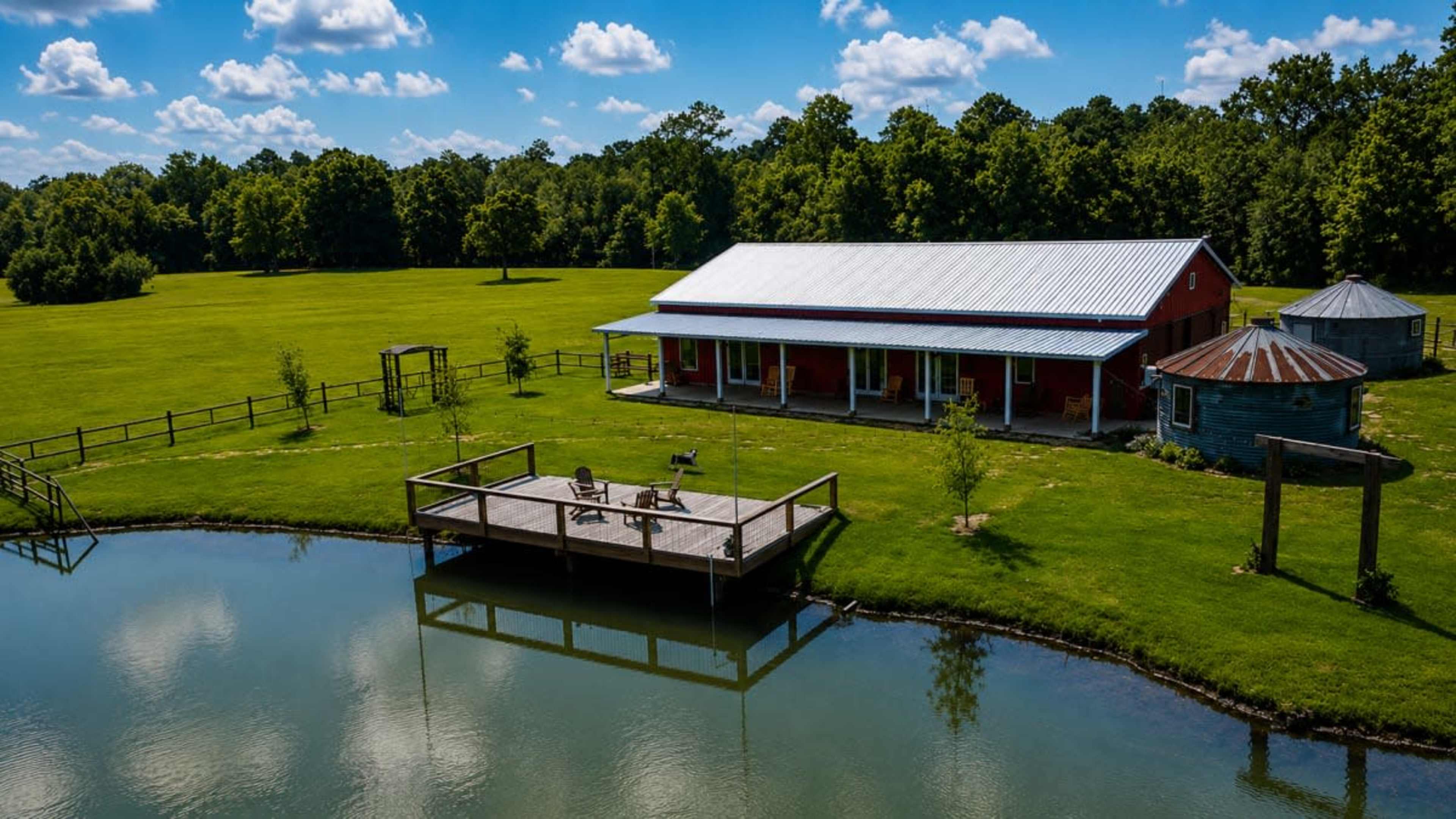 A red barn-style building sits next to a pond, with a wooden deck and chairs overlooking the water, surrounded by a green field and trees.