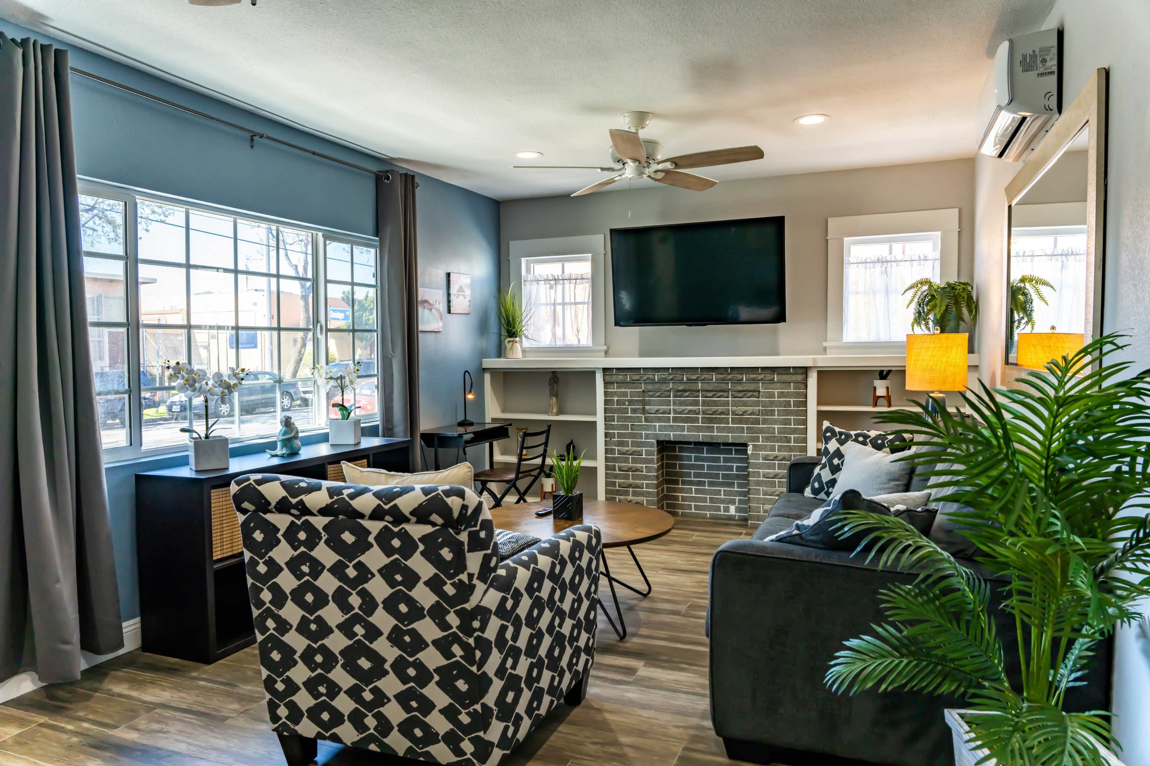 The image shows a modern living room with a gray color scheme, featuring a sectional sofa, an armchair, a coffee table, a flat-screen TV mounted above a brick fireplace, and large windows allowing natural light to enter.