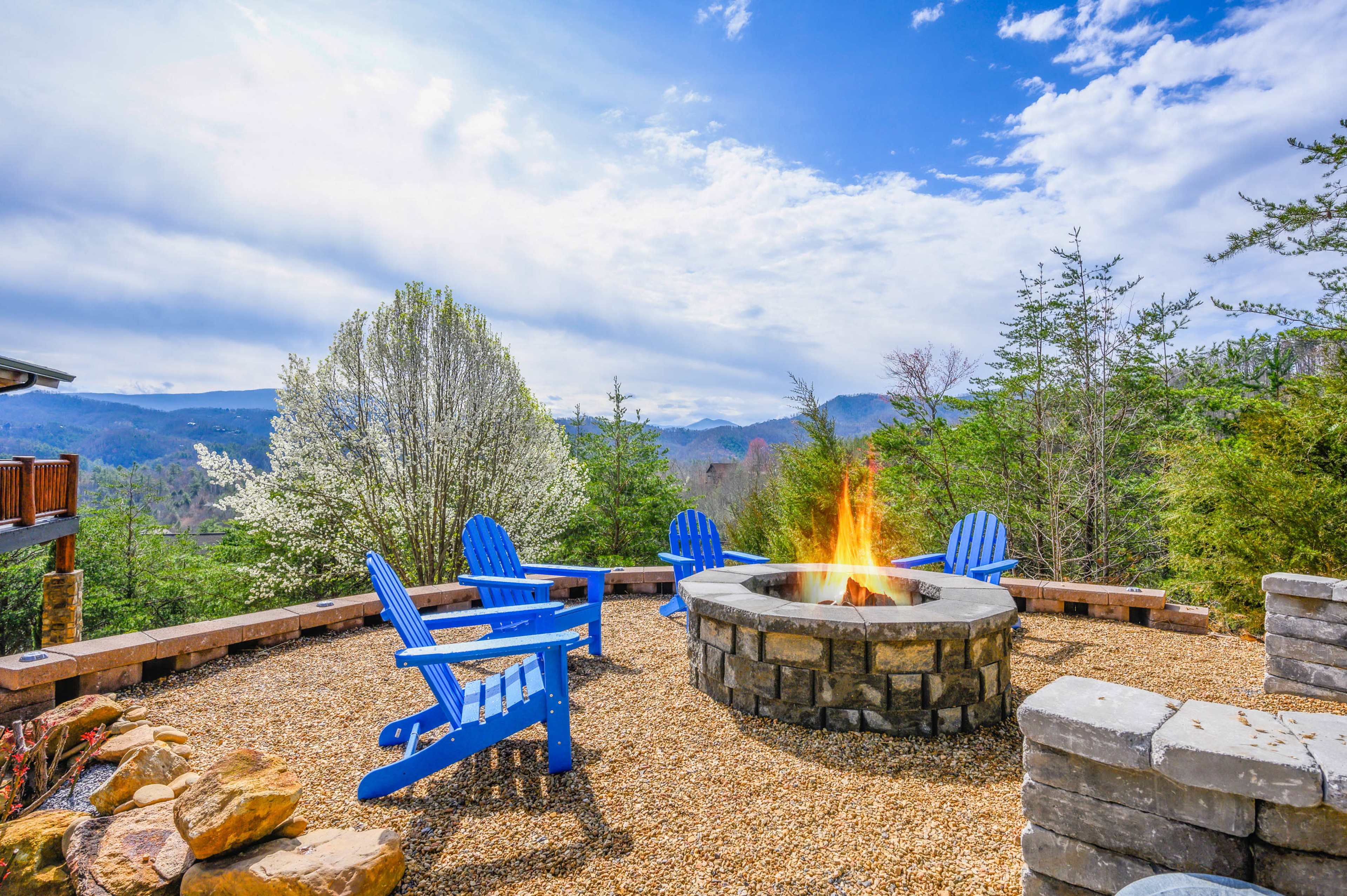 A stone fire pit surrounded by blue Adirondack chairs sits on a gravel patio, with trees and mountains visible in the background under a partly cloudy sky.