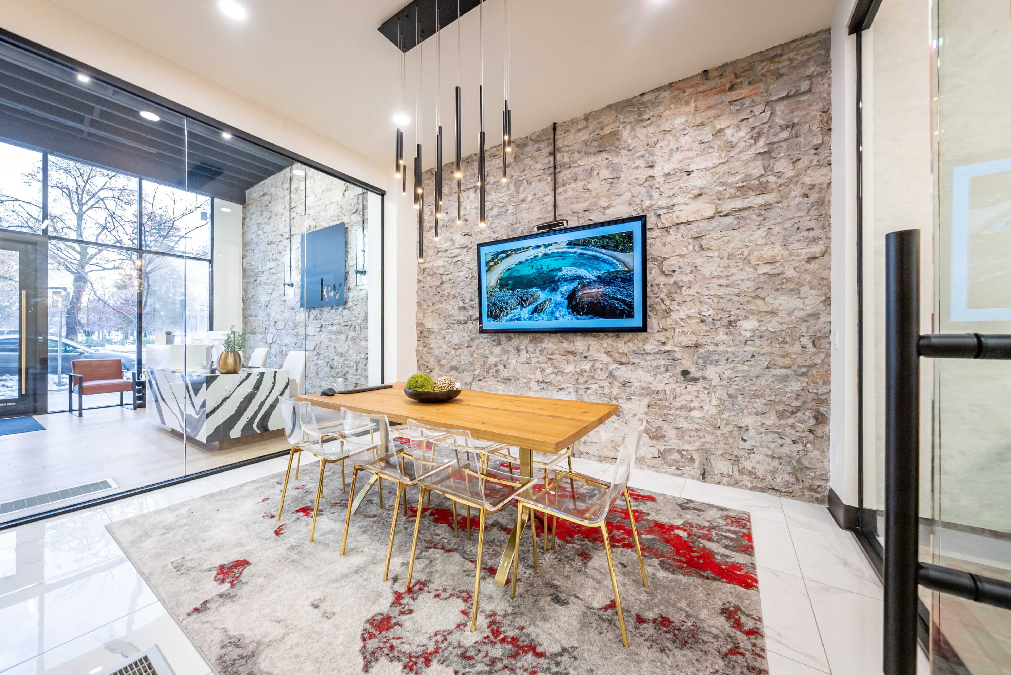 A modern dining area with a wooden table, clear chairs, a large television mounted on a stone wall, and a decorative rug on the floor.