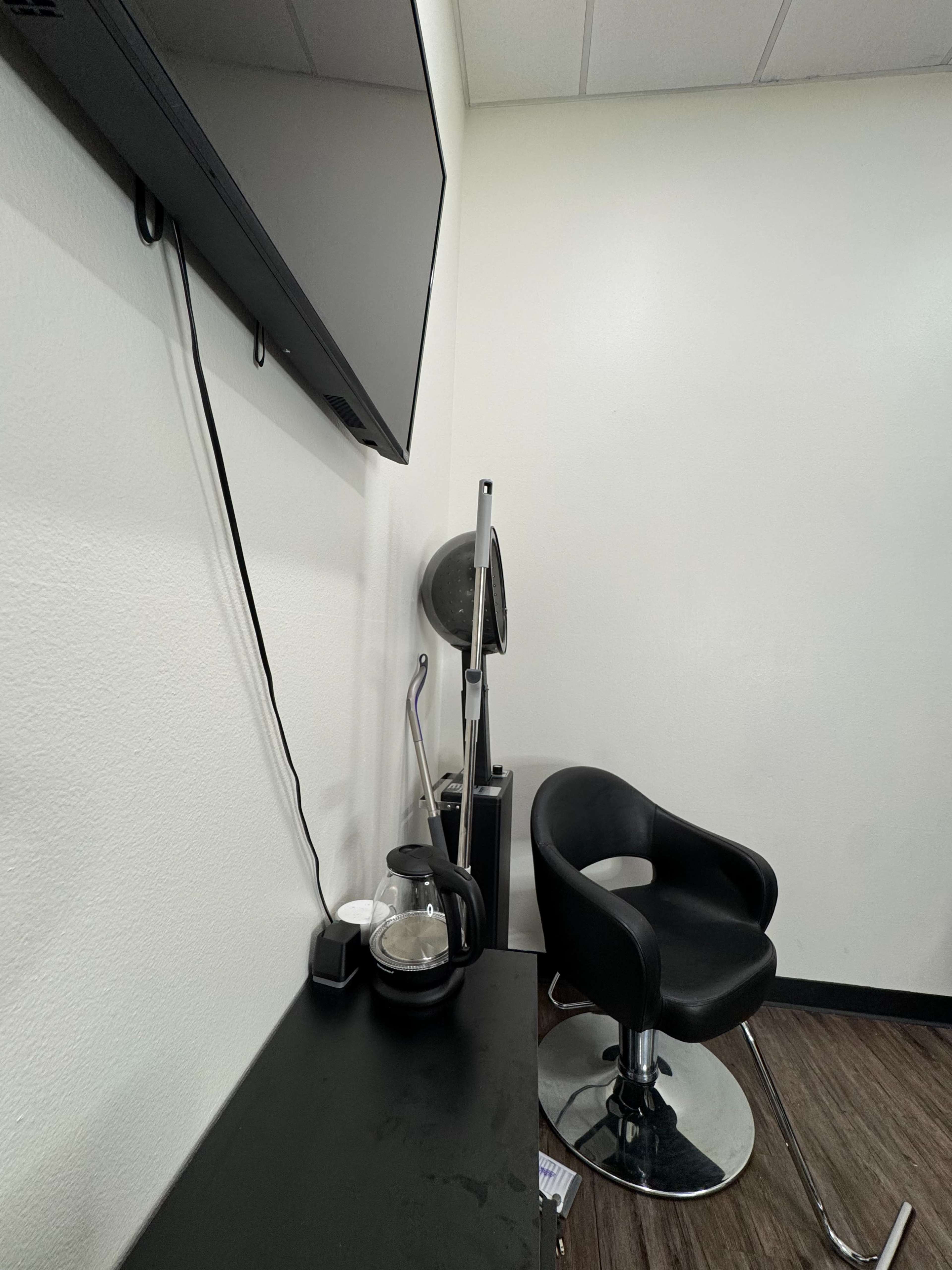 The image shows a minimalistic salon space with a black chair, a wall-mounted TV, and a small table containing a kettle.