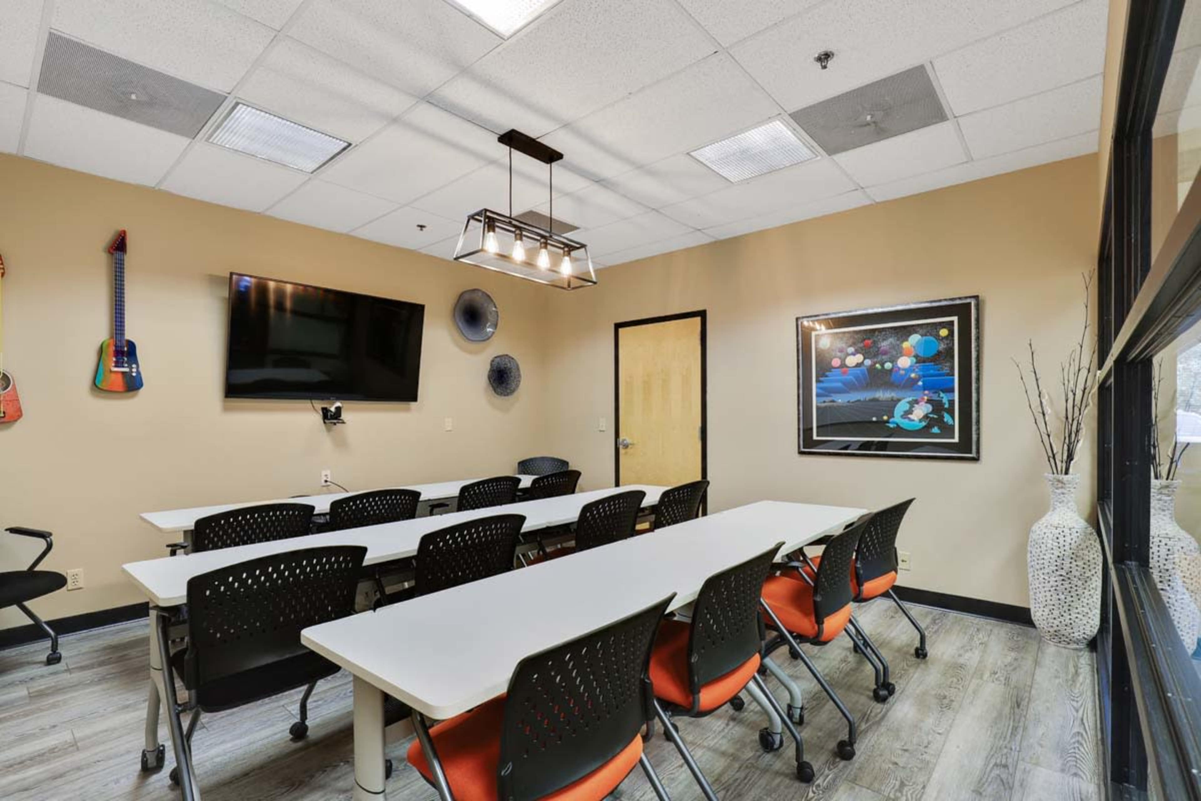 A well-lit meeting room features a long table with rolling chairs, a wall-mounted TV, and musical instruments displayed on the walls.