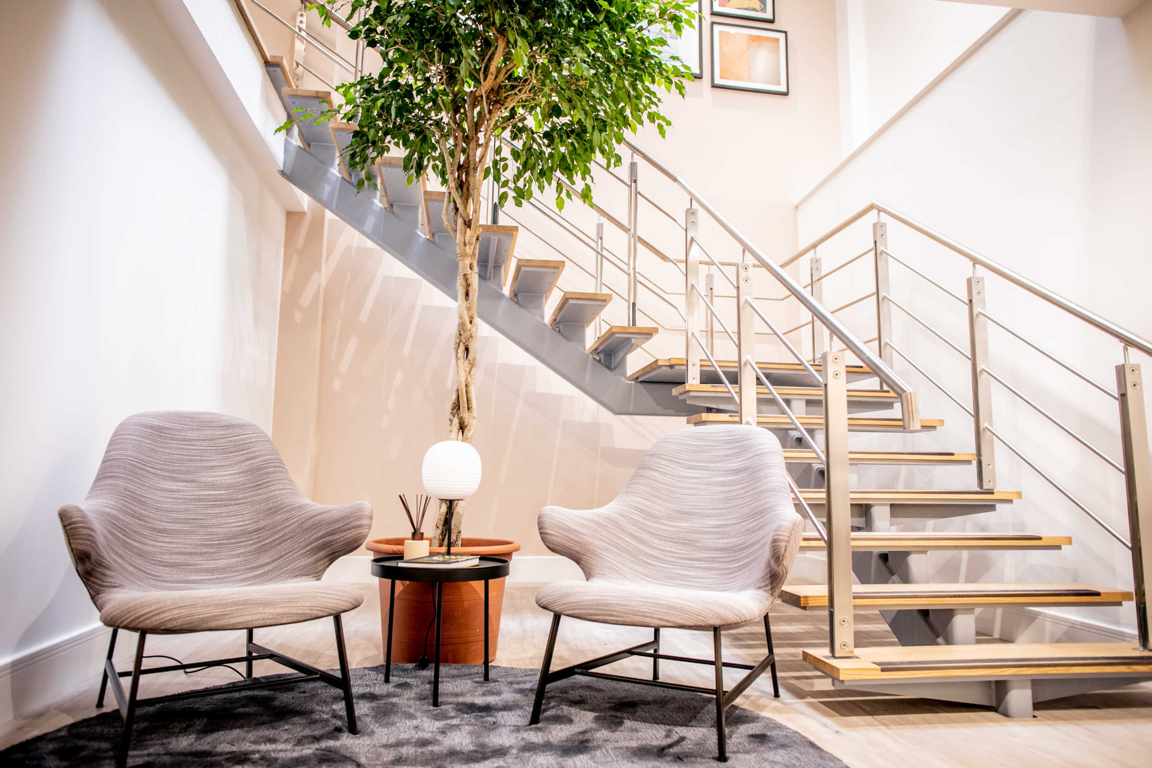 A cozy seating area with two gray armchairs, a small table with a lamp, and a potted tree beside a staircase.