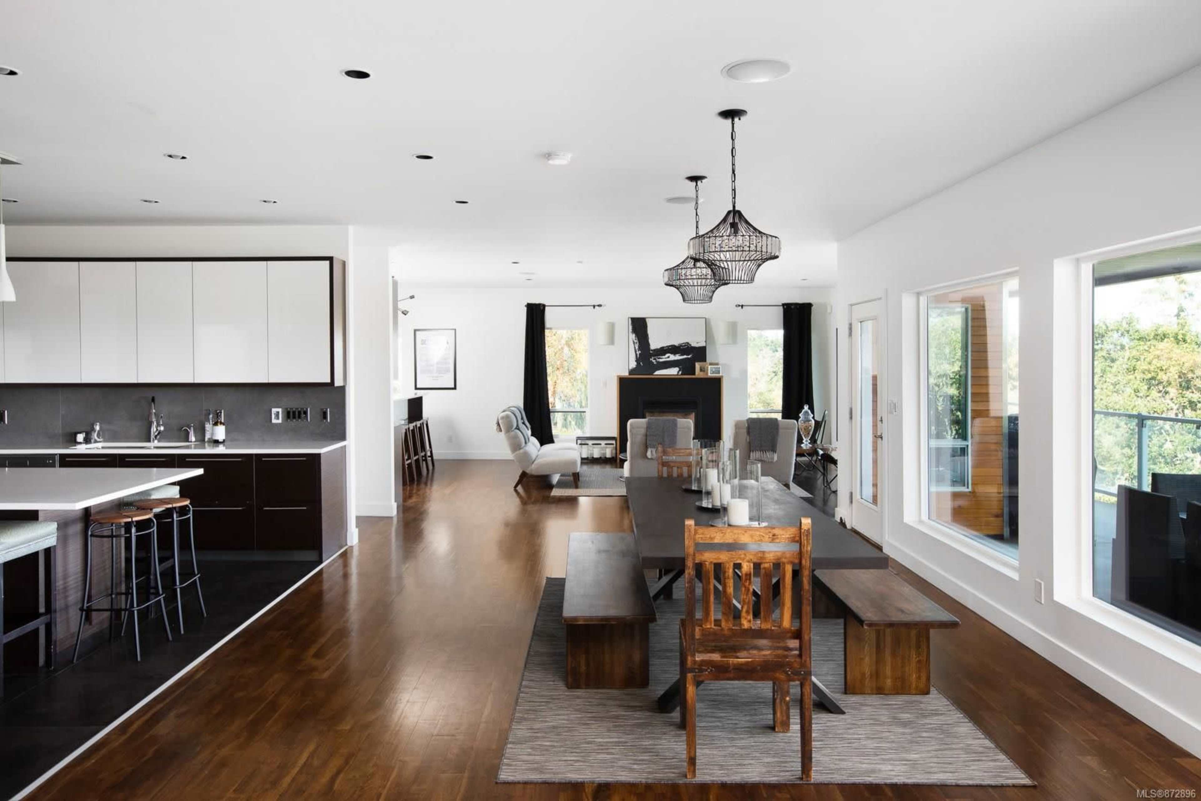 A modern open-concept living and dining area with a long wooden table, black kitchen cabinets, and large windows overlooking greenery.