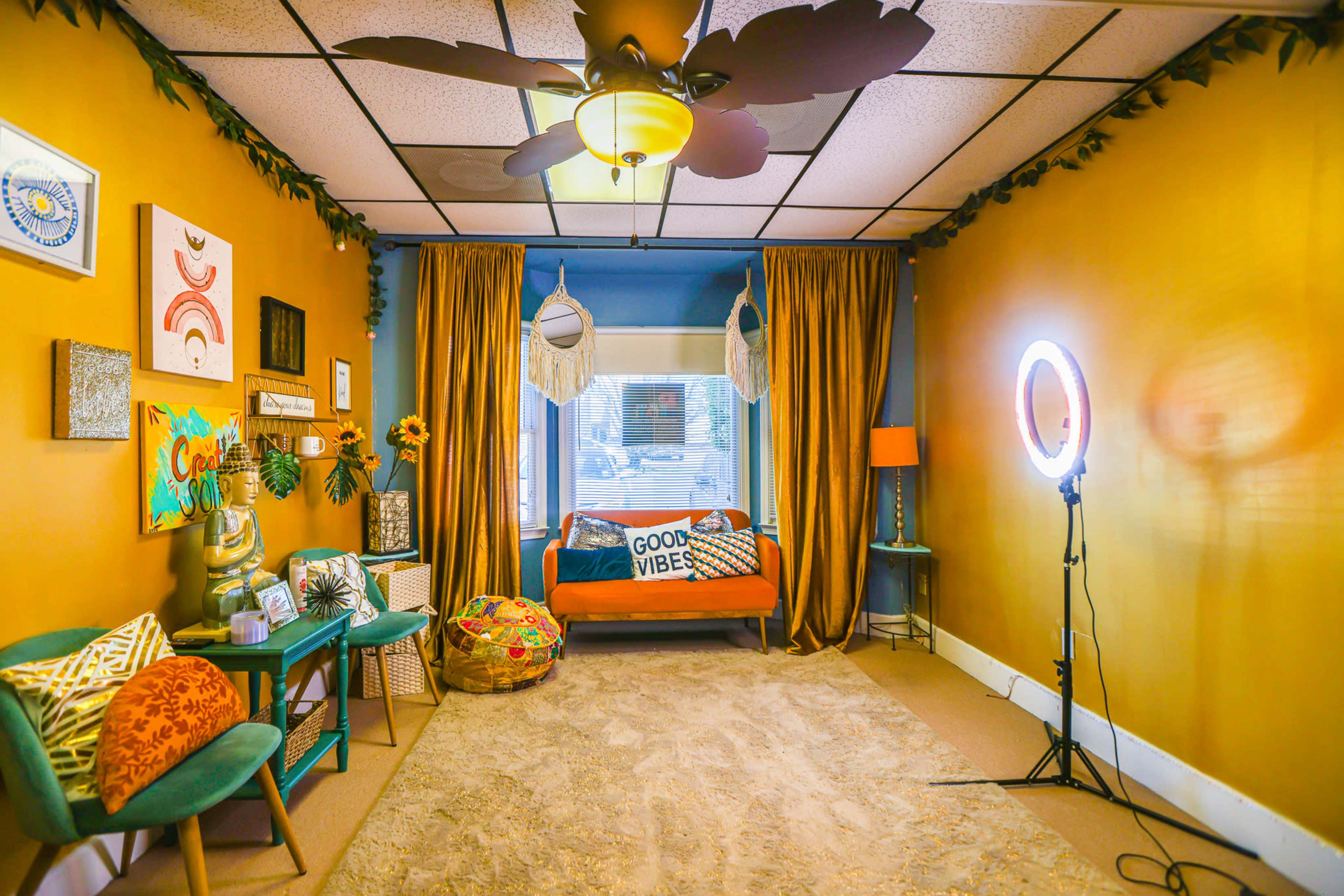 The image shows a brightly colored room with an orange couch, decorative pillows, a large ring light, curtains, and wall art, all arranged around a cozy area rug.