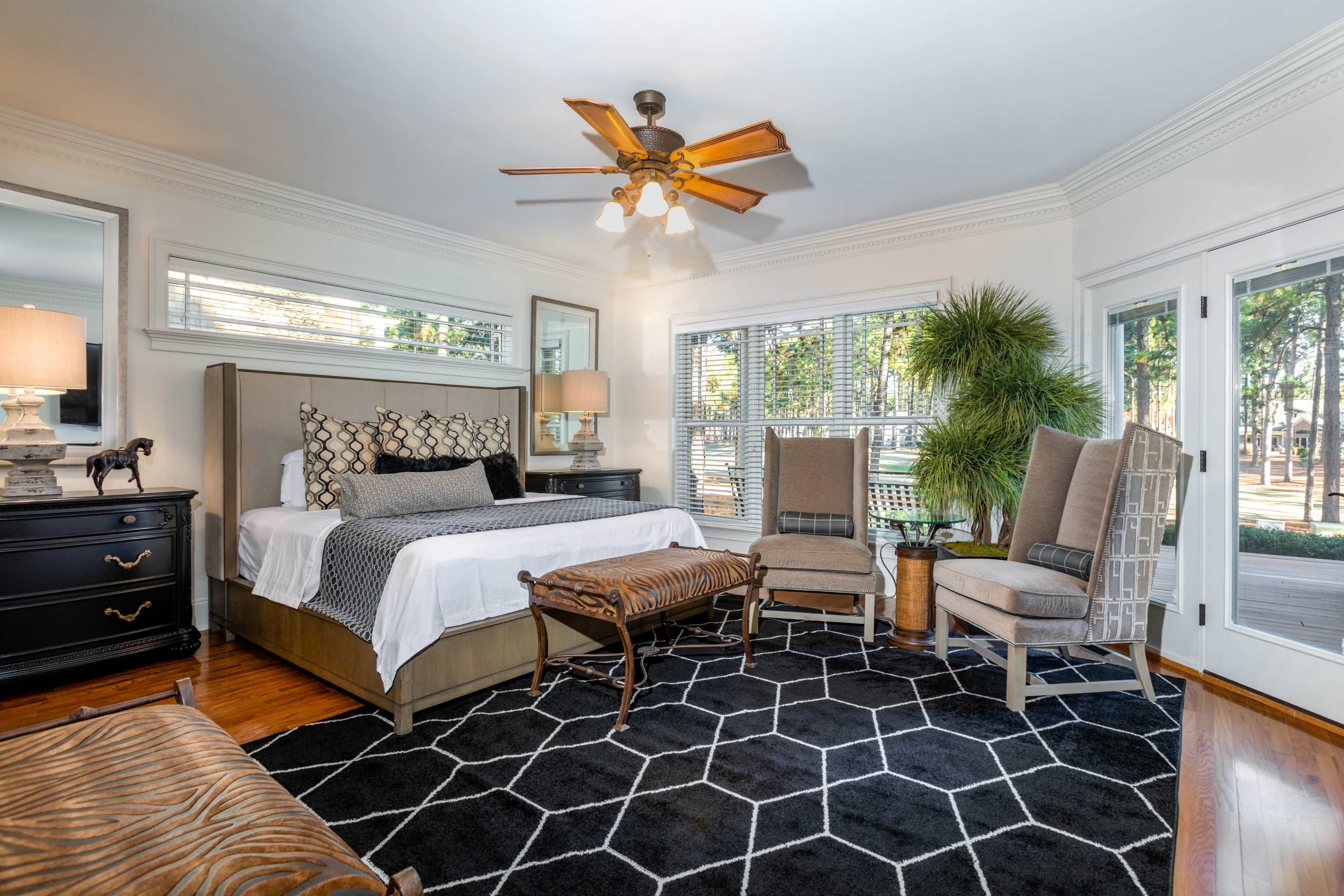 The bedroom features a large bed, two armchairs, a patterned rug, and large windows overlooking a garden.
