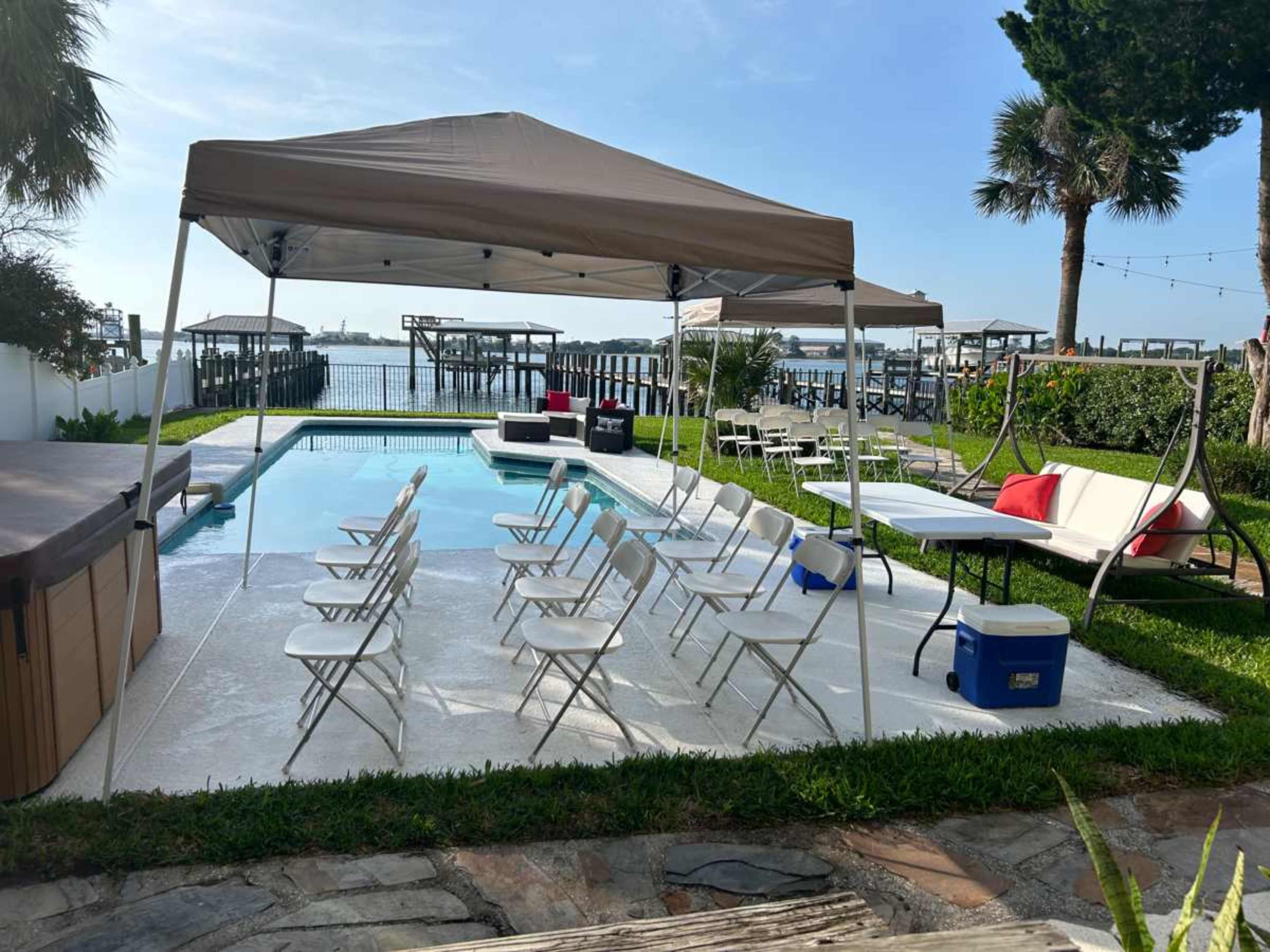 A tent is set up beside a pool surrounded by chairs and tables, with a view of a waterfront dock and palm trees in the background.