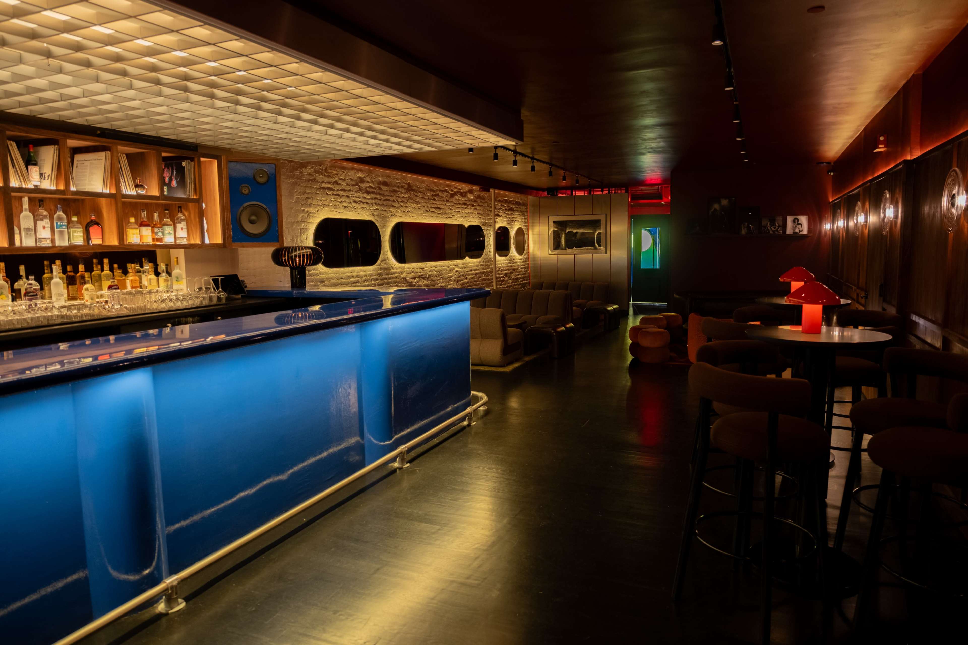 The image shows a dimly lit bar interior with a blue illuminated countertop, shelves stocked with liquor, and a seating area featuring modern furniture.