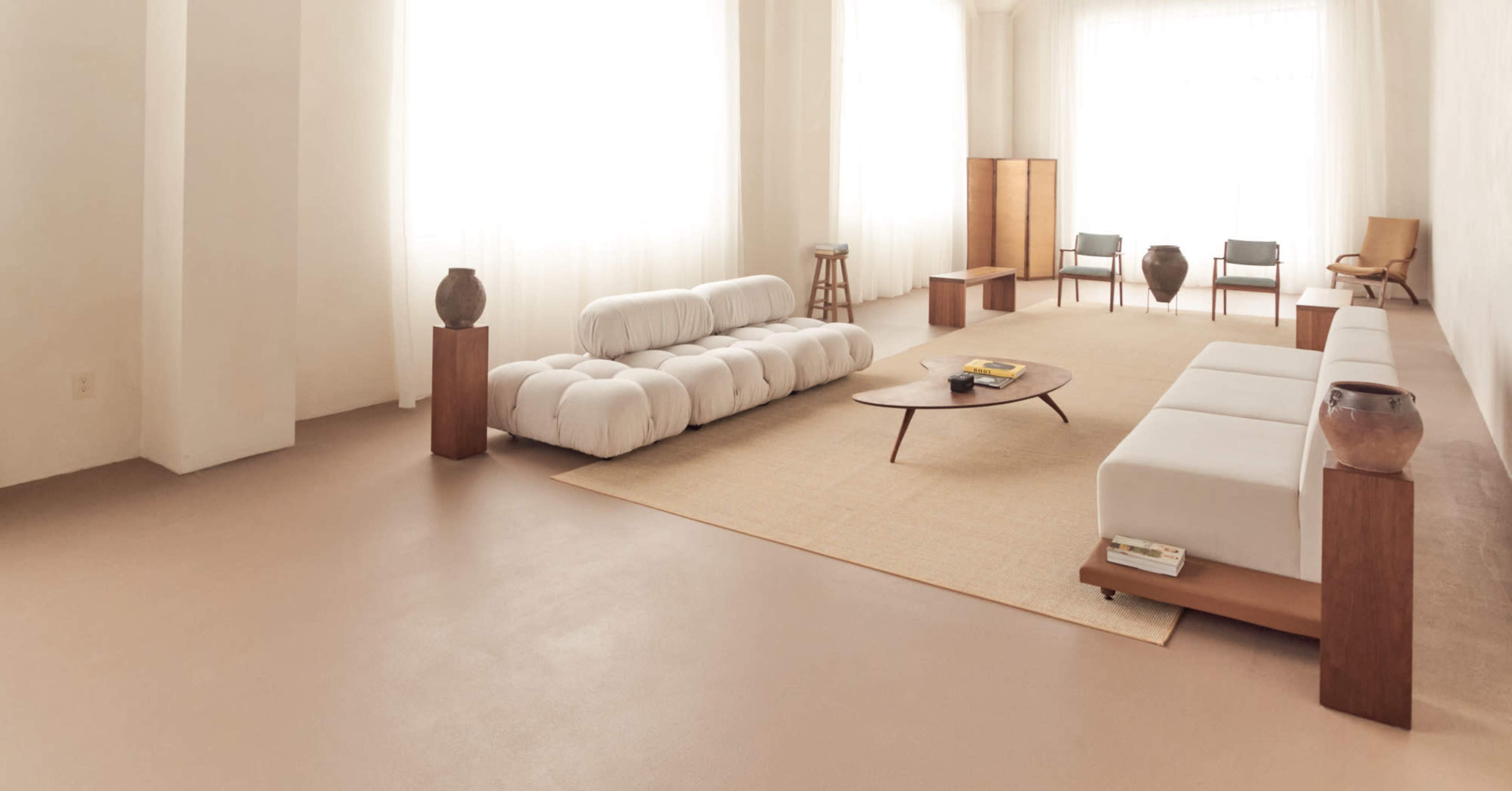 The image shows a modern, minimalist living room featuring light-colored walls, a large area rug, a low coffee table, and various seating options, all illuminated by natural light from large windows.