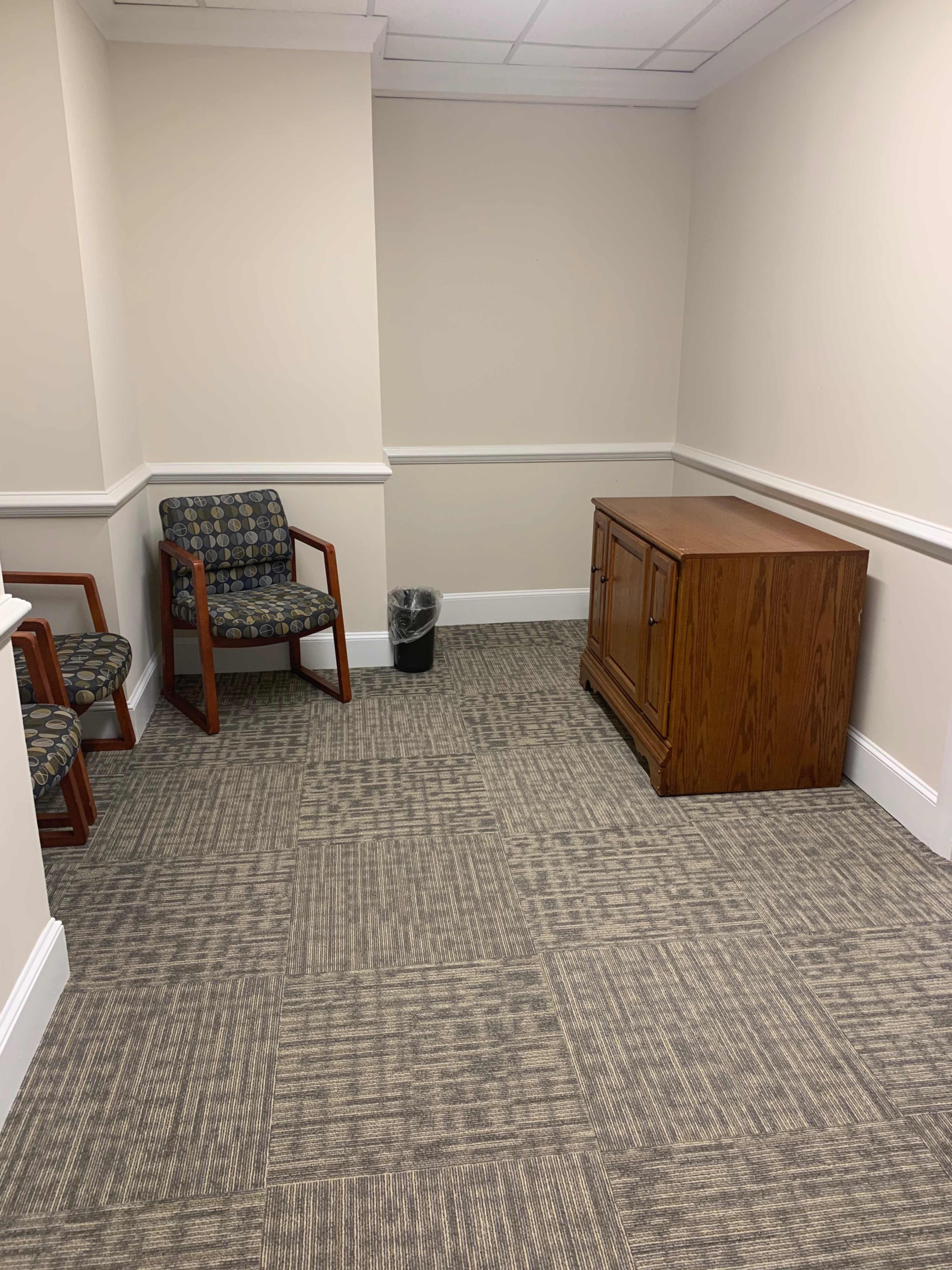 The image shows a small corner of an office with two chairs, a wooden cabinet, and a trash can on the carpeted floor.