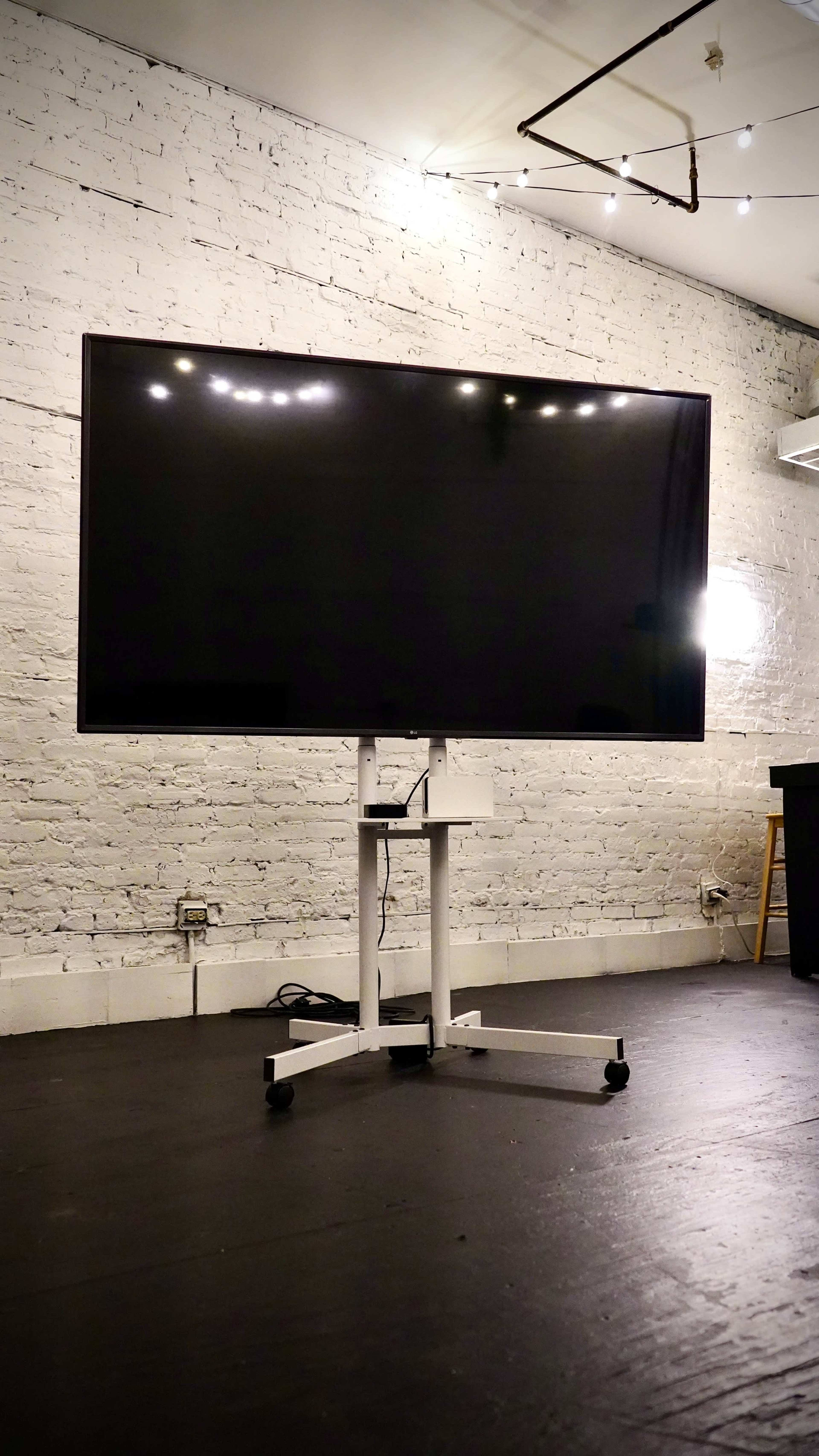 A large, blank screen monitor on a wheeled stand is positioned in front of a textured white brick wall.