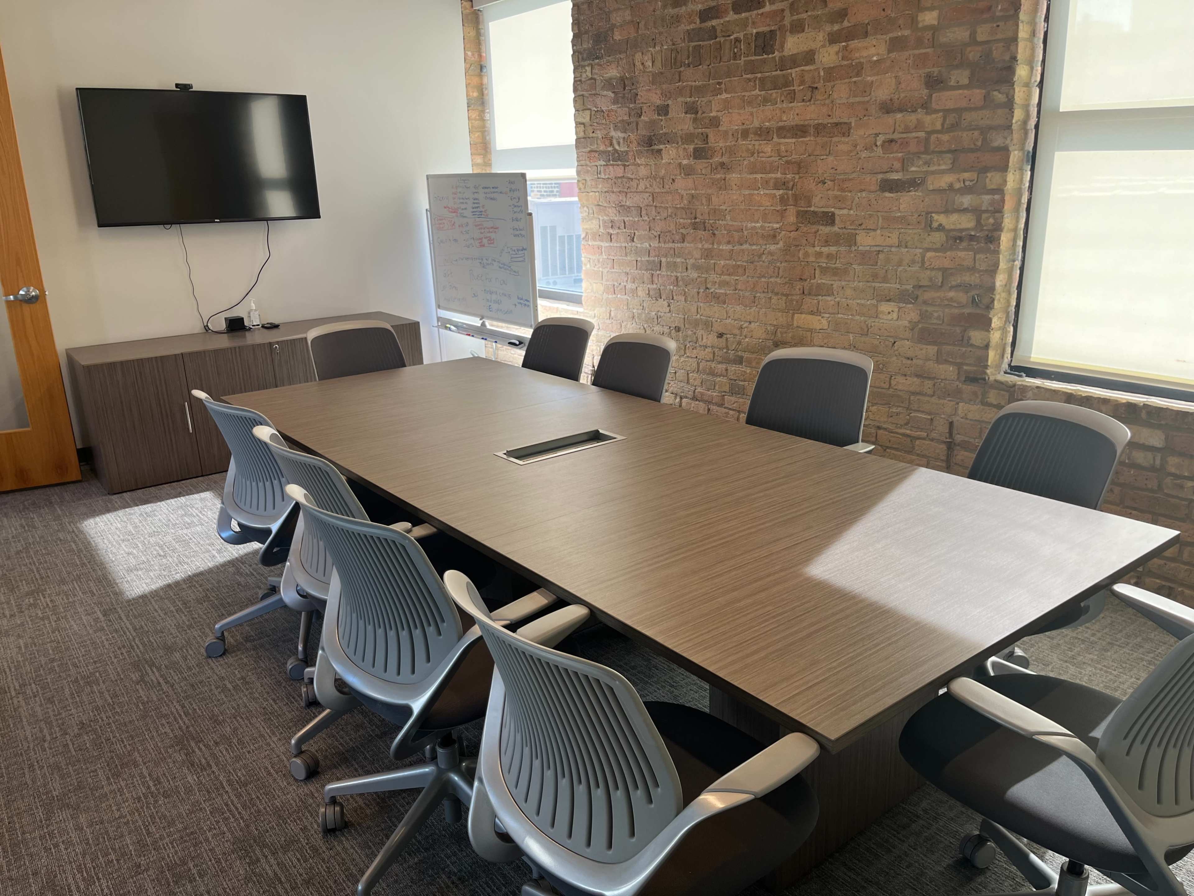 A modern conference room features a large rectangular wooden table surrounded by ergonomic chairs, with a television mounted on a wall and a whiteboard nearby.
