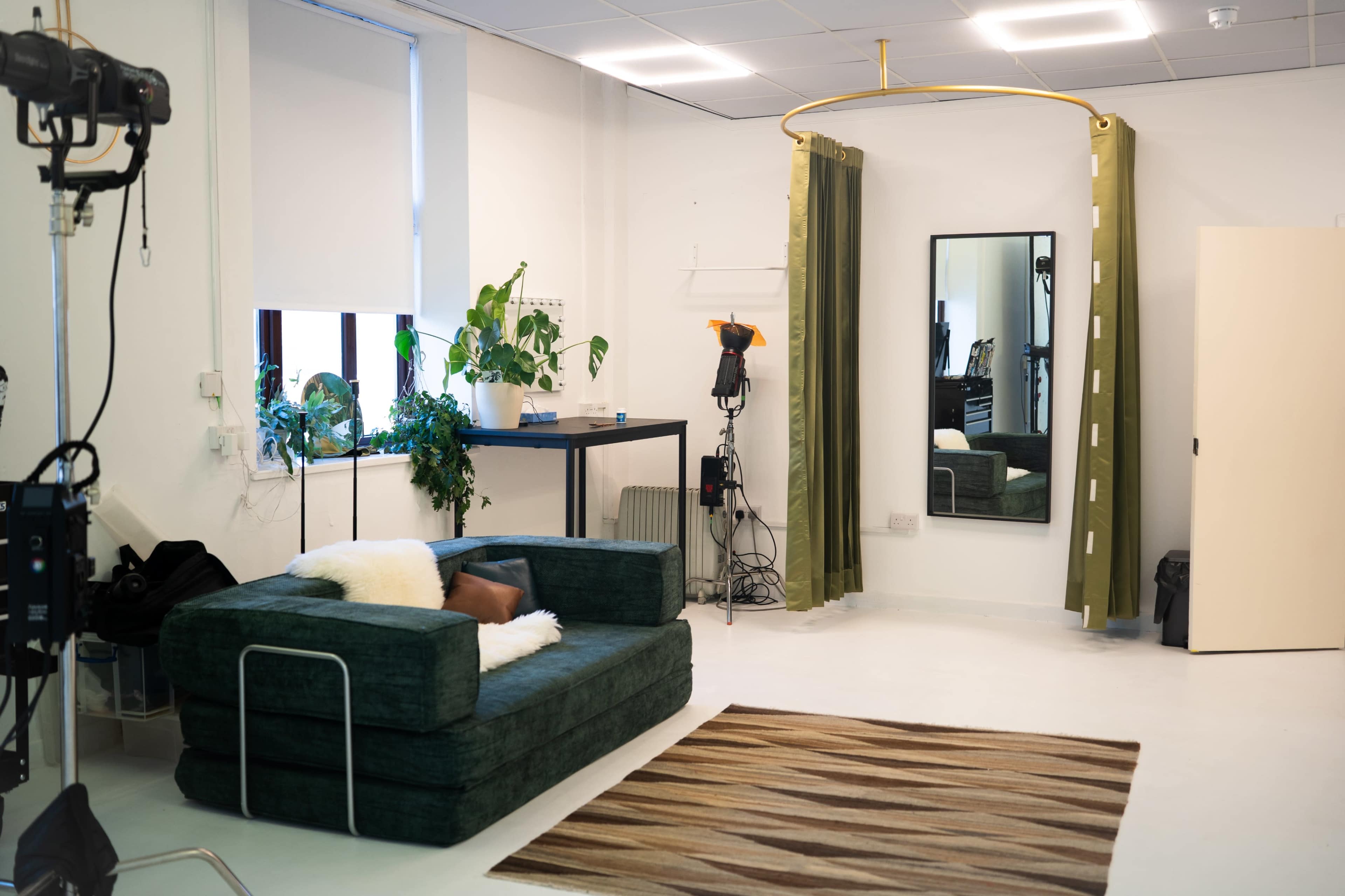 The image shows a modern, minimalist studio space featuring a green sofa, a rug, a mirror, and plants, with a backdrop setup for photography.