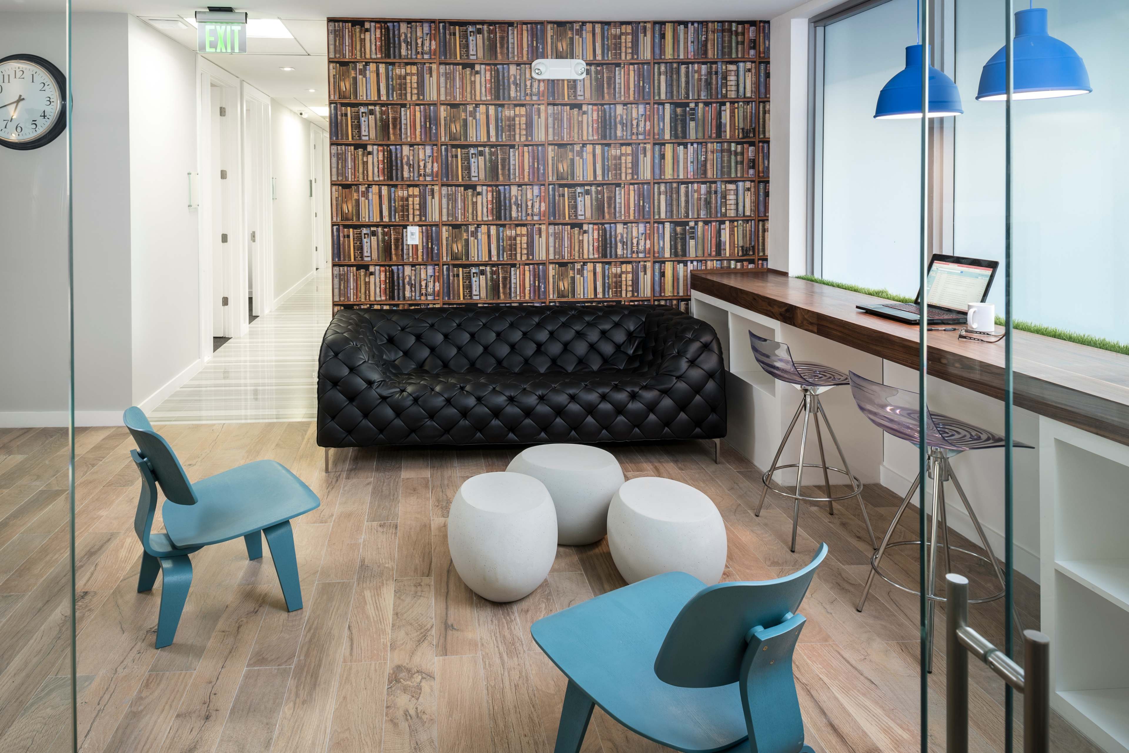 The image shows a modern lounge area with a black tufted sofa, circular white stools, and blue chairs, alongside a wall designed to look like a row of bookshelves.