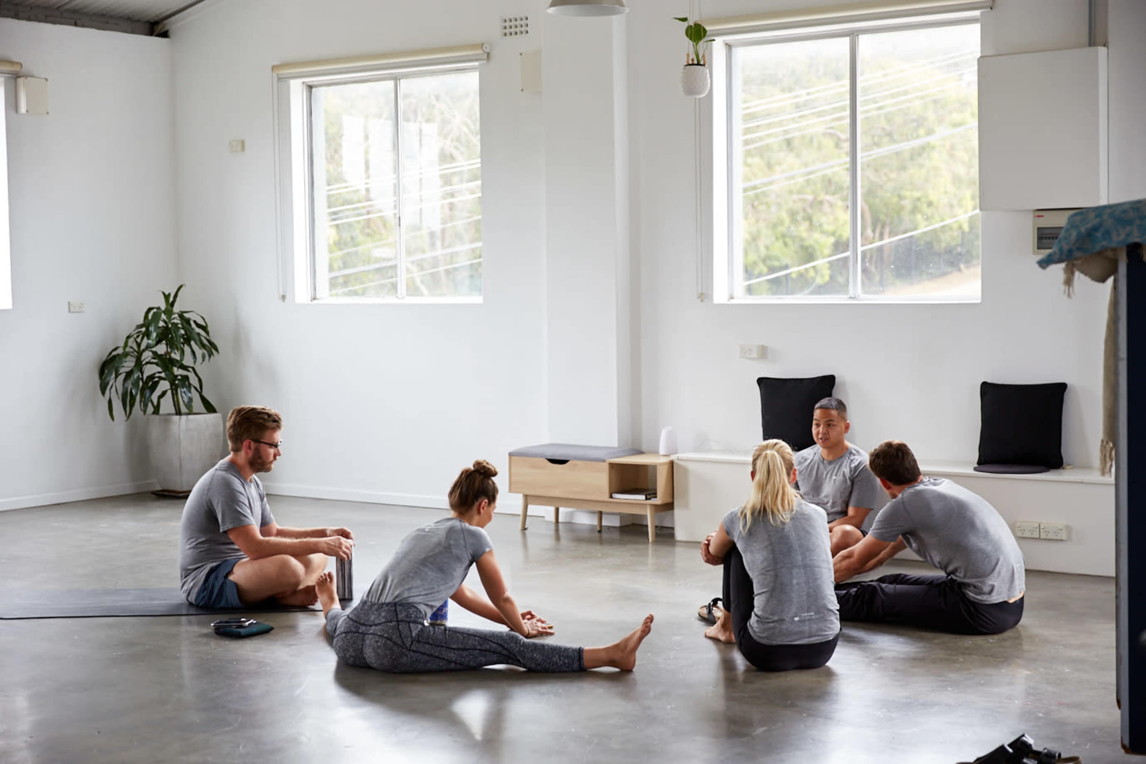 A group of five people sits on the floor in a light-filled room, engaging in conversation while some stretch on yoga mats.