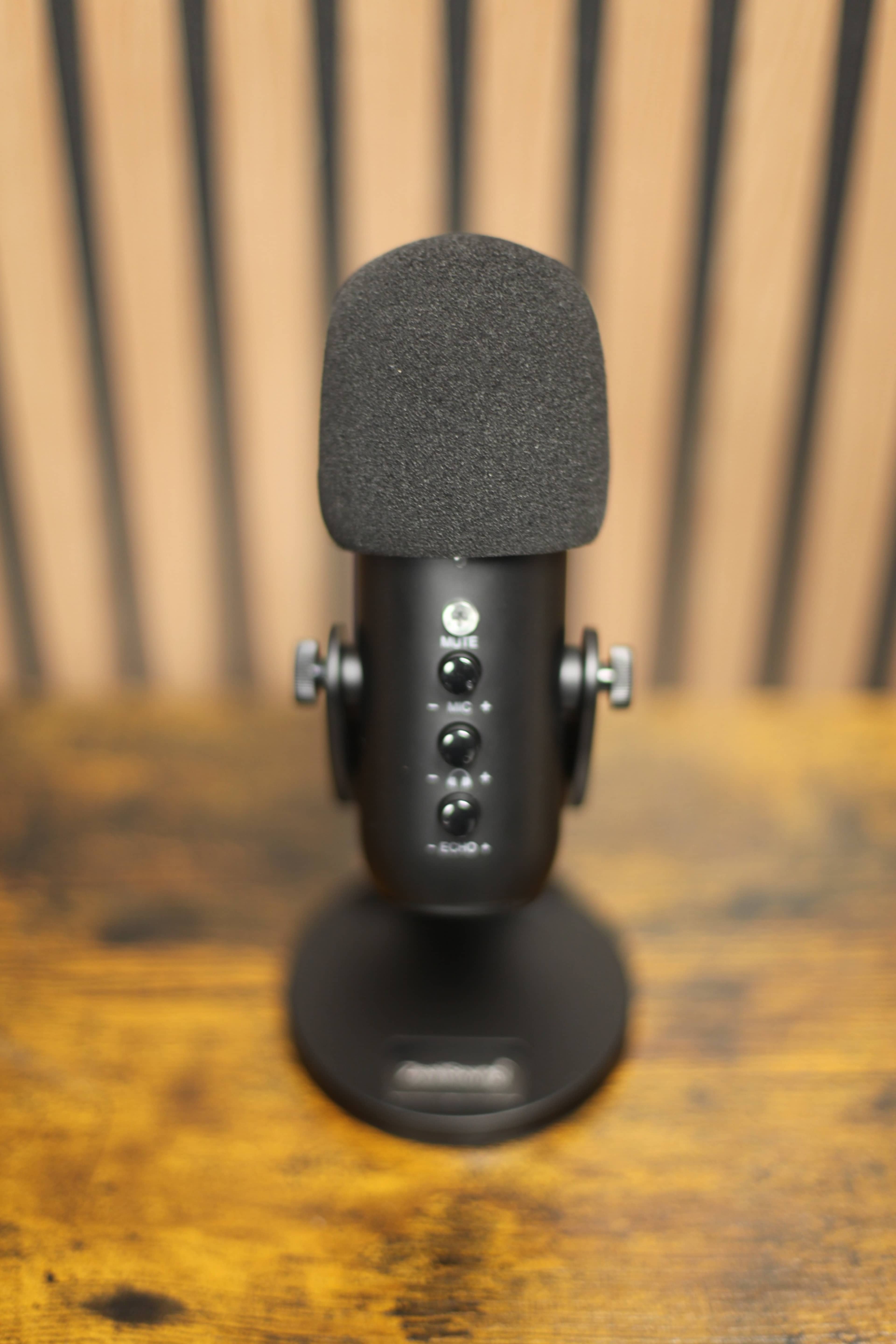 A black microphone with a foam cover stands upright on a wooden surface, featuring several control buttons on its front.