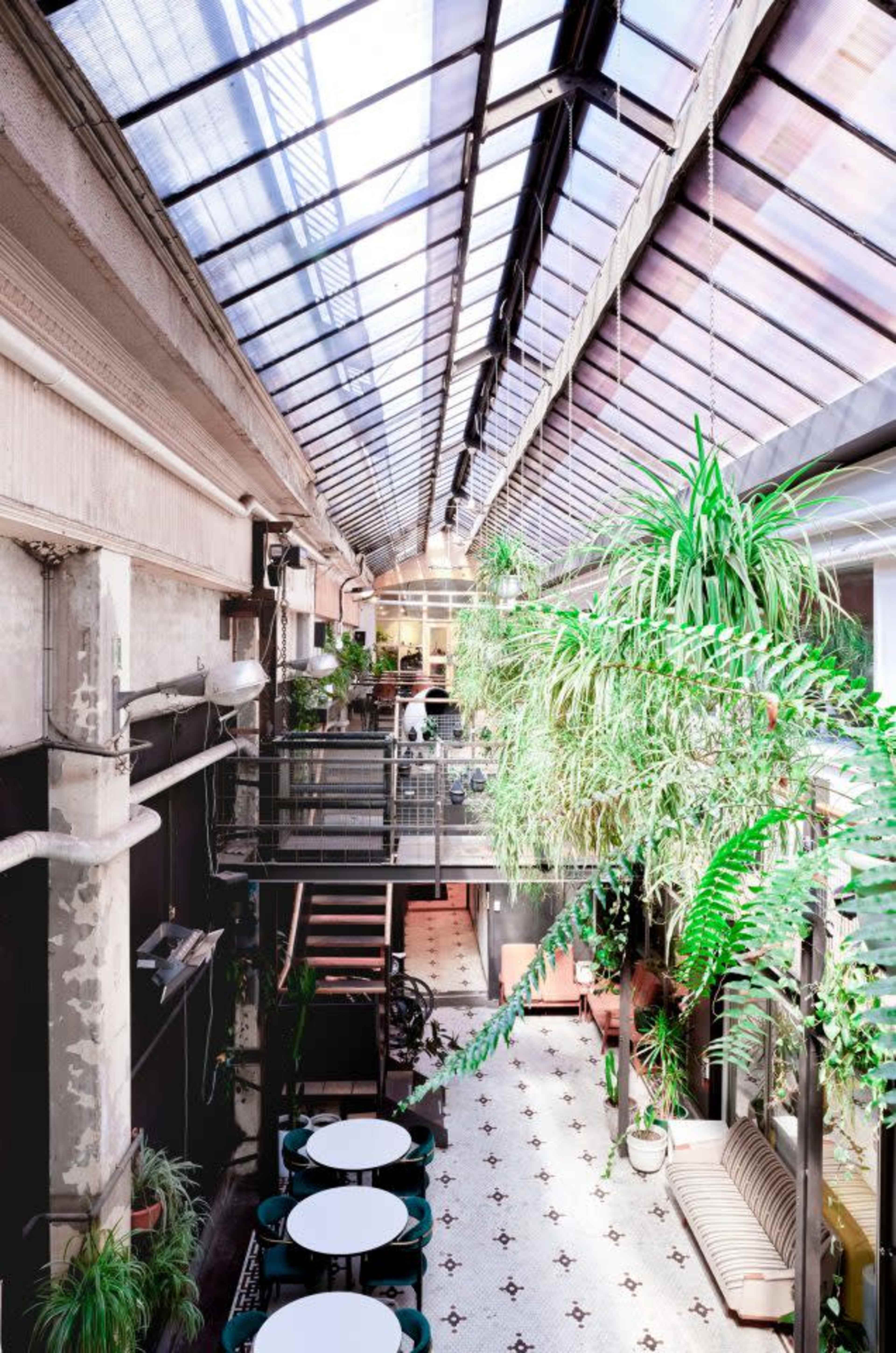A long, airy interior space with a glass roof, featuring greenery, seating areas, and a patterned tiled floor.