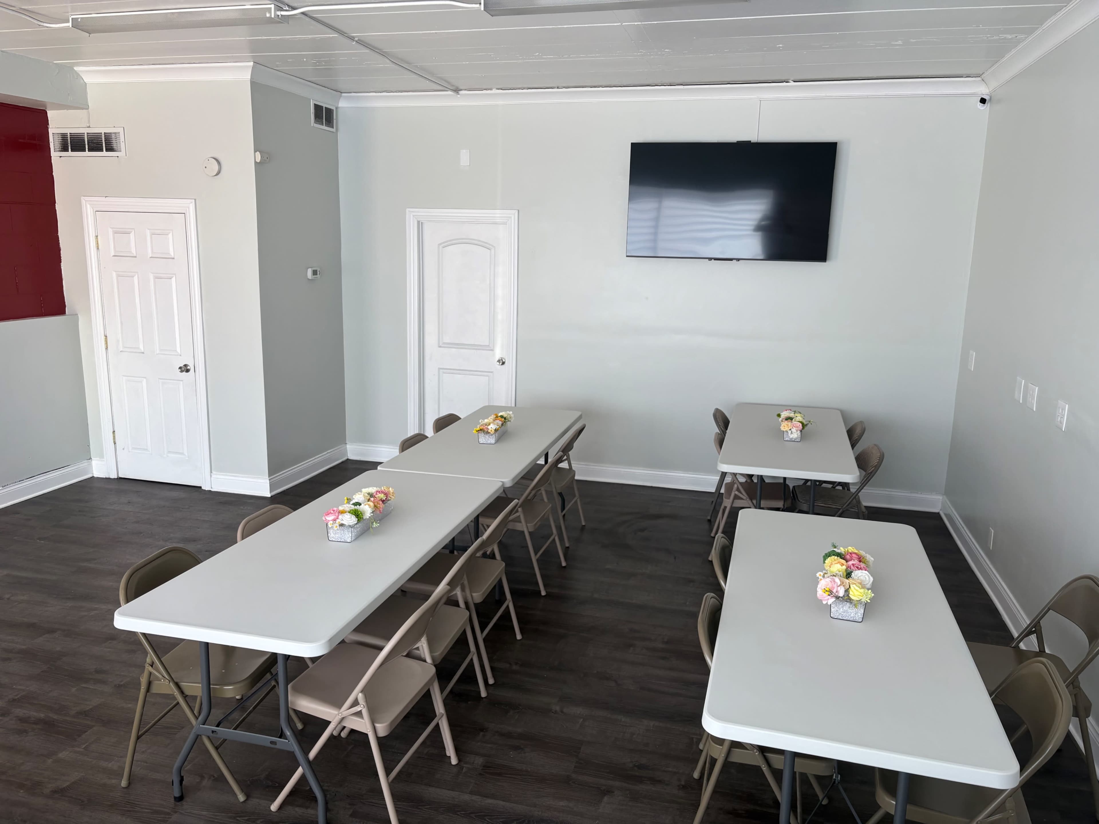 The image shows a brightly lit room with four tables arranged neatly, each adorned with a small floral centerpiece, and a mounted television on the wall.