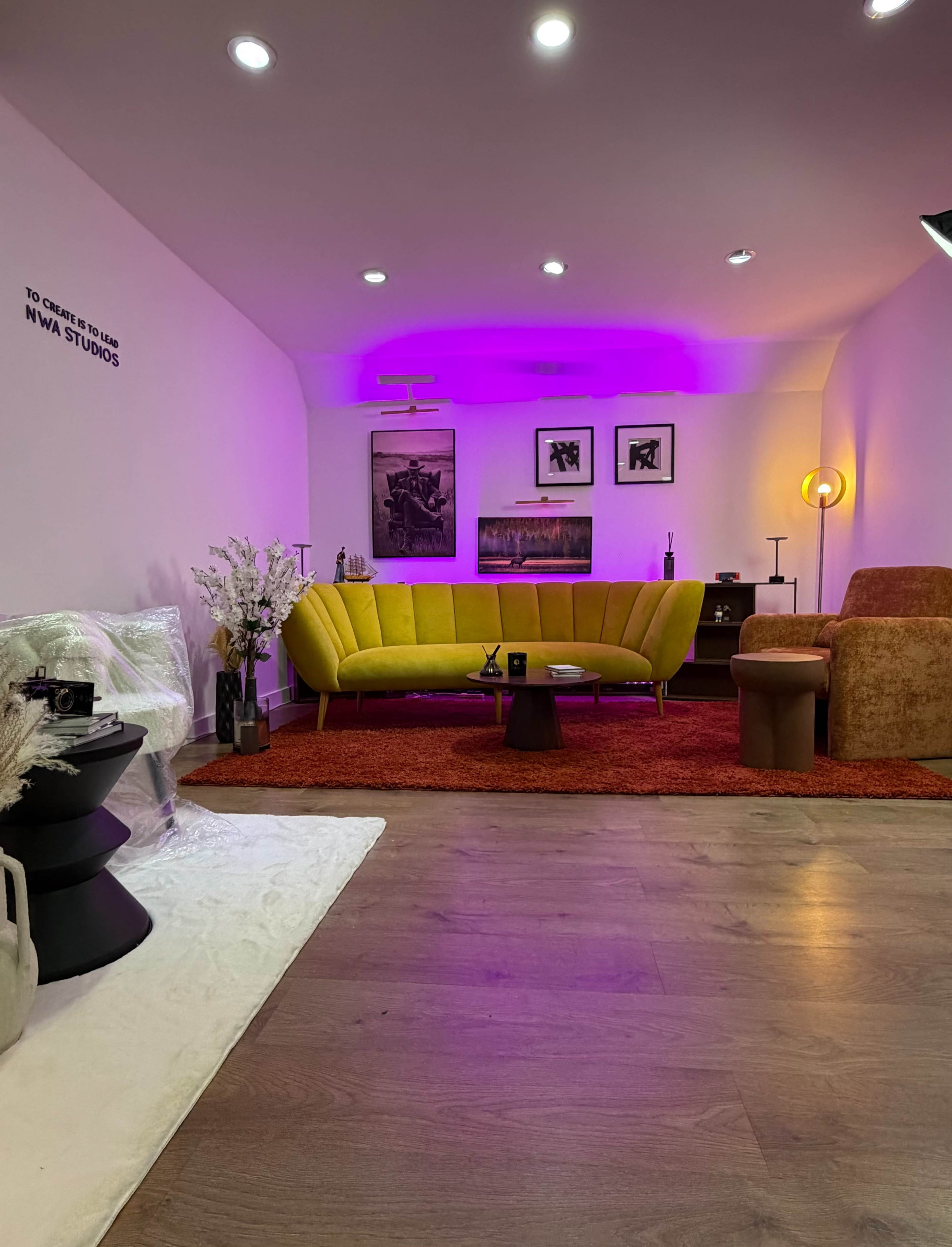The image shows a modern living room featuring yellow and brown couches, artwork on the walls, and purple ambient lighting.