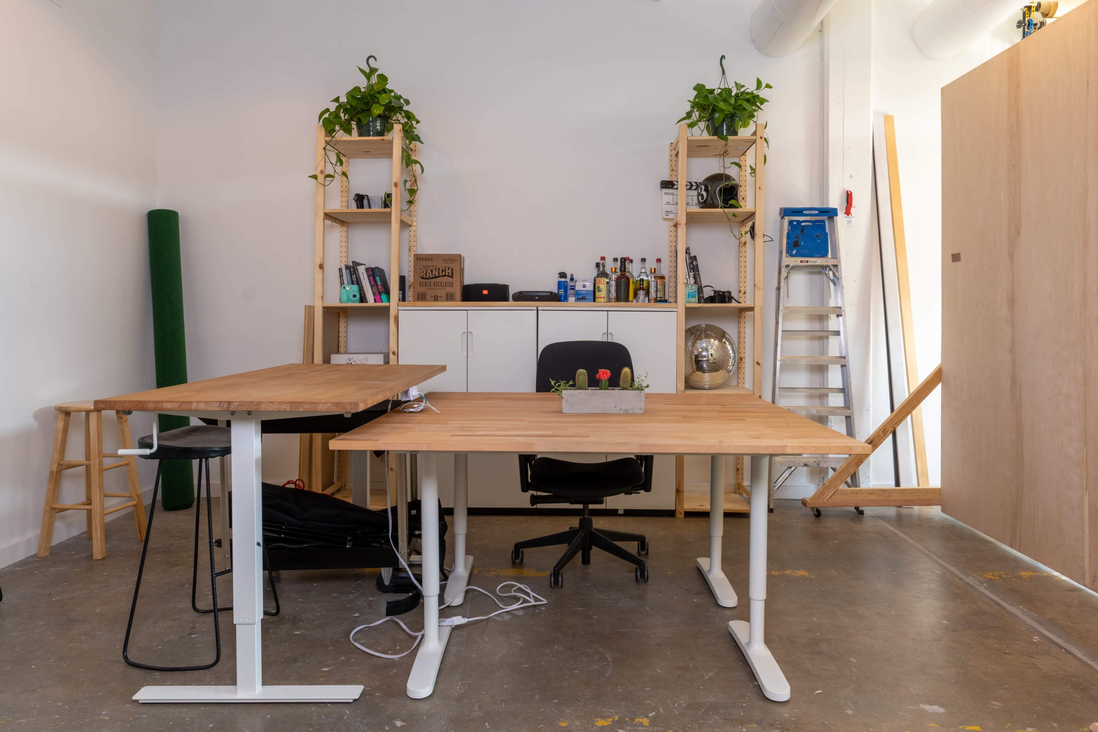 A modern office space features two wooden desks, a black office chair, shelving with various items, and greenery, set against a minimalistic backdrop.
