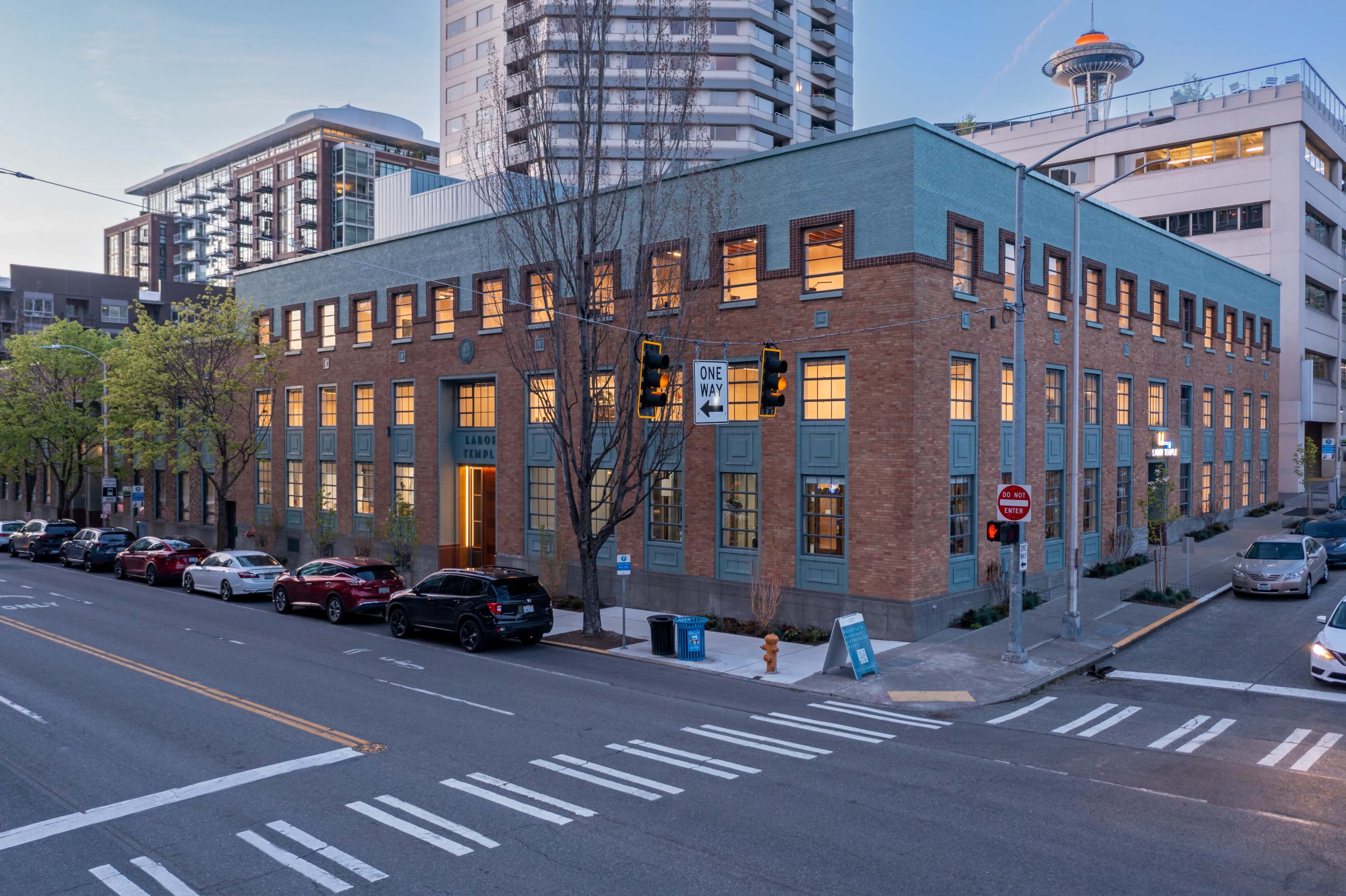 A modern brick building with large windows located at a street intersection in an urban area, with nearby high-rise buildings and a prominent space needle in the background.