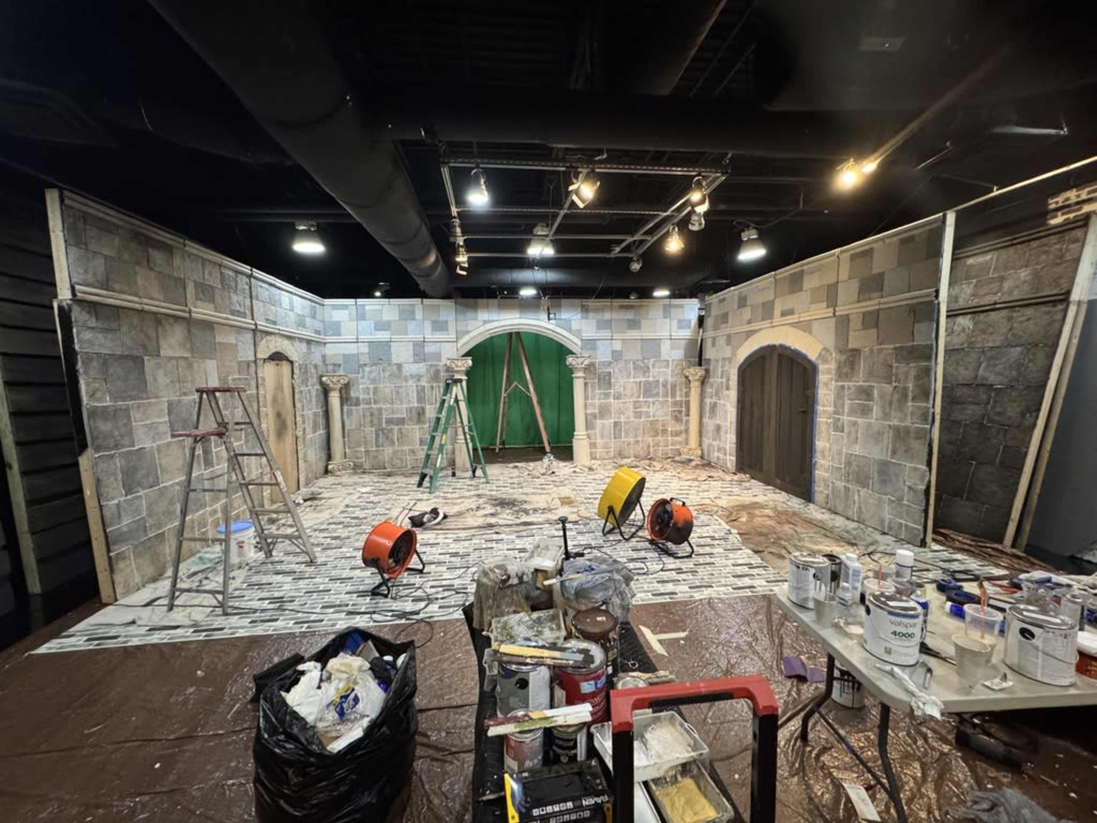 The image shows an unfinished interior scene resembling a medieval setting, with stone walls, various construction equipment, and paint supplies scattered across the floor.