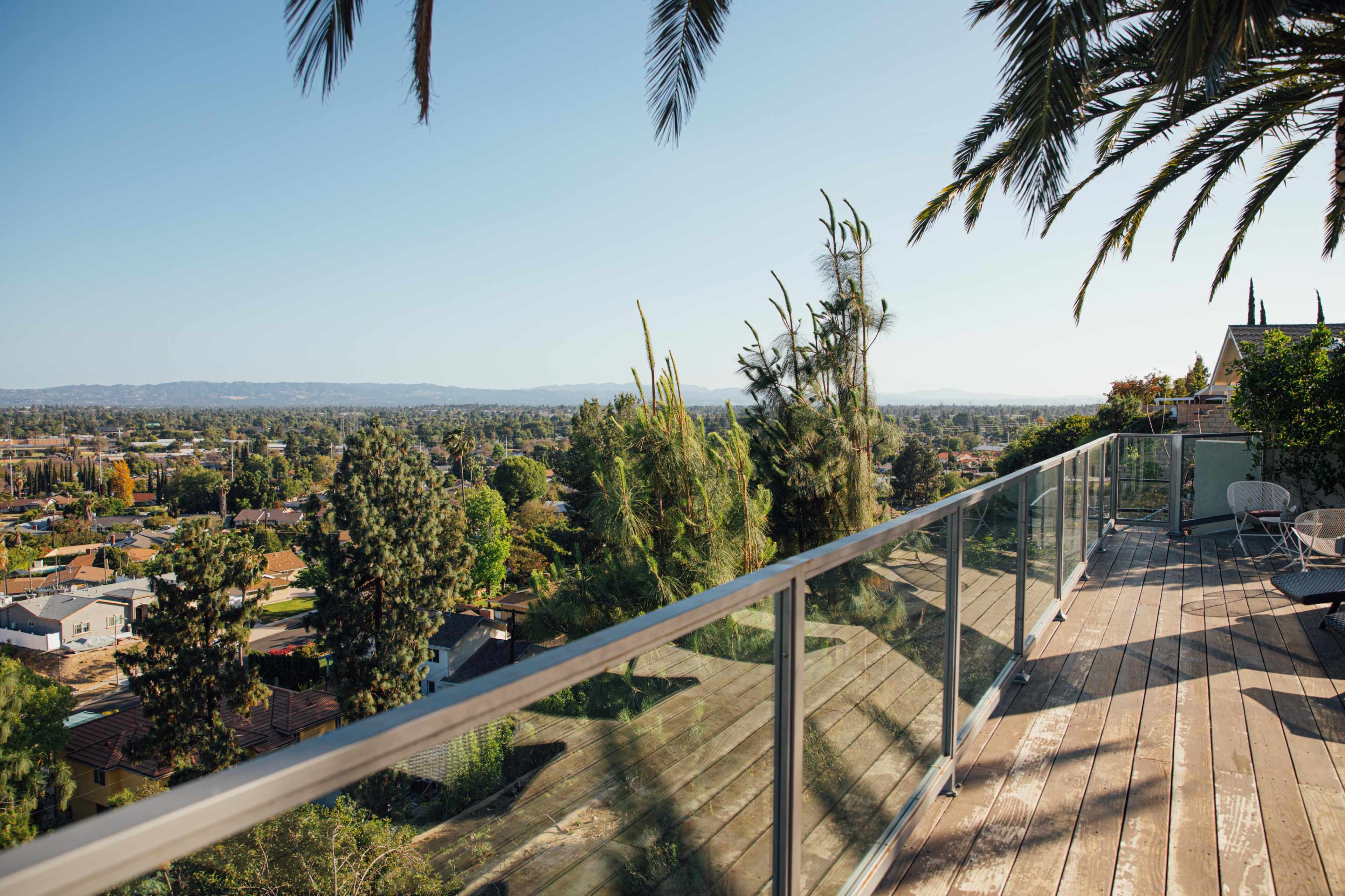 A balcony with a glass railing overlooks a sprawling urban landscape with trees and distant mountains.
