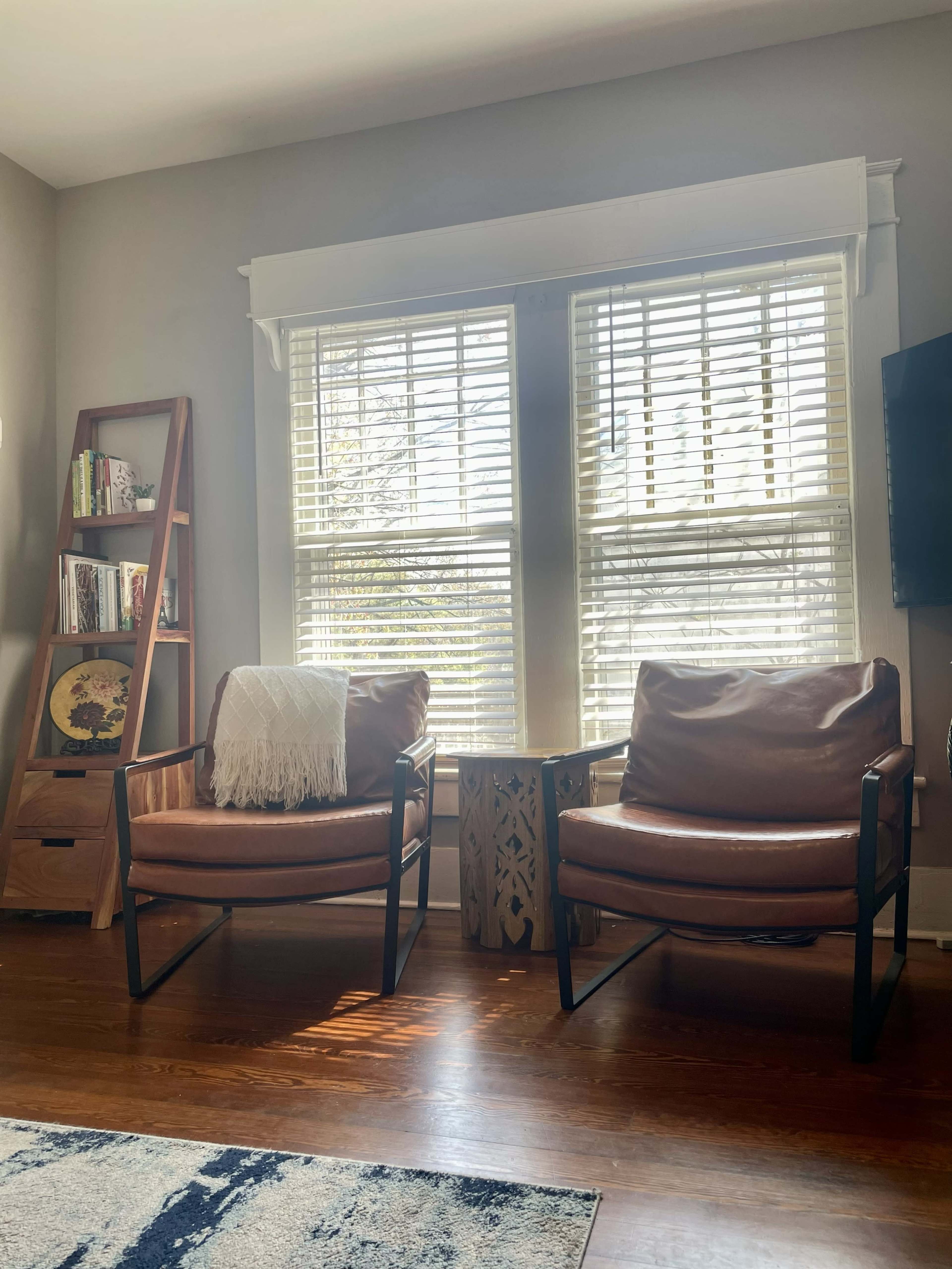 The image shows a cozy living room with two brown leather chairs facing a wooden side table, flanked by a bookshelf and large windows with blinds allowing natural light to enter.
