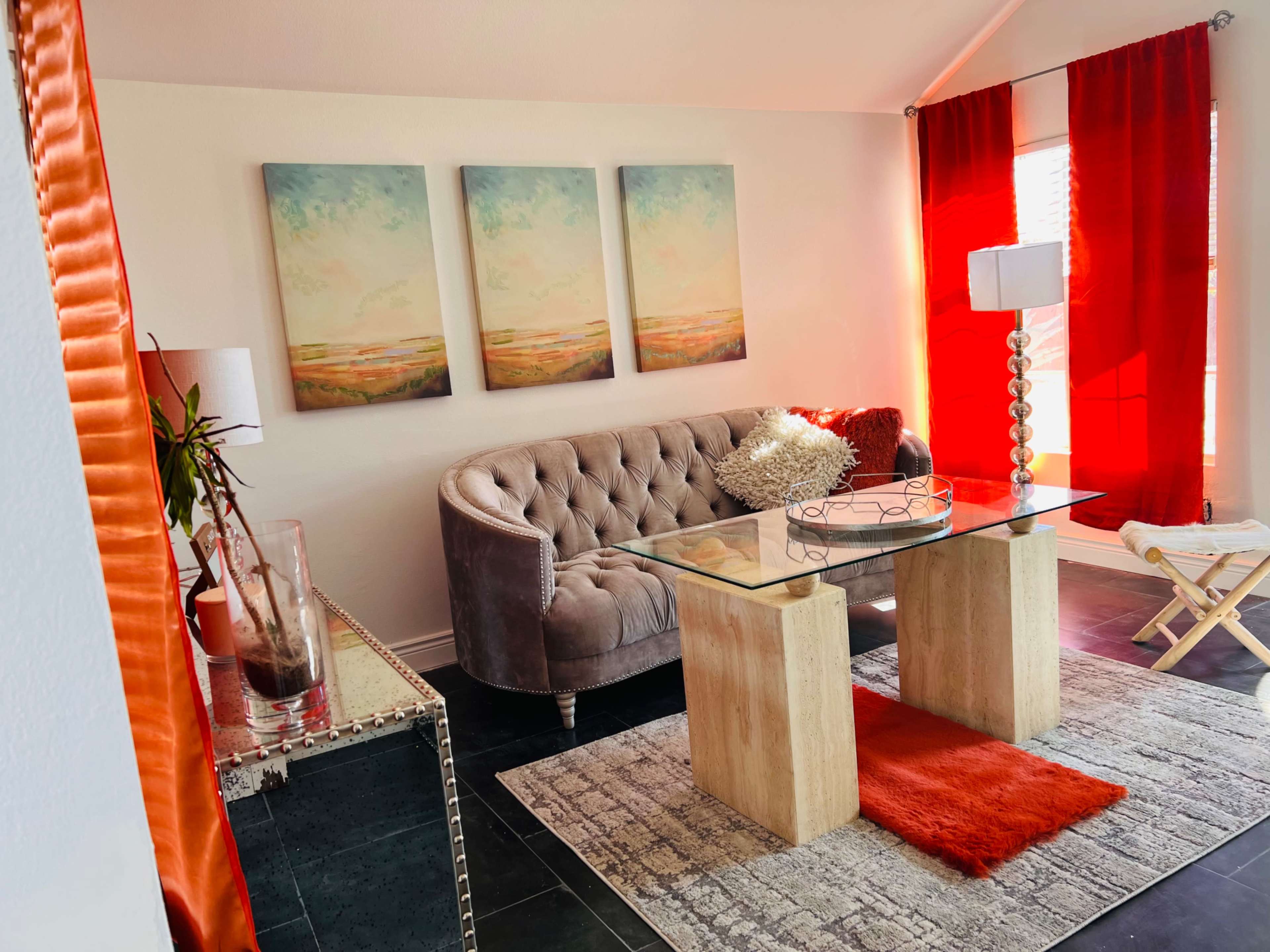 A modern living room with a tufted gray sofa, a glass coffee table, and bright red curtains, complemented by three landscape paintings on the wall.