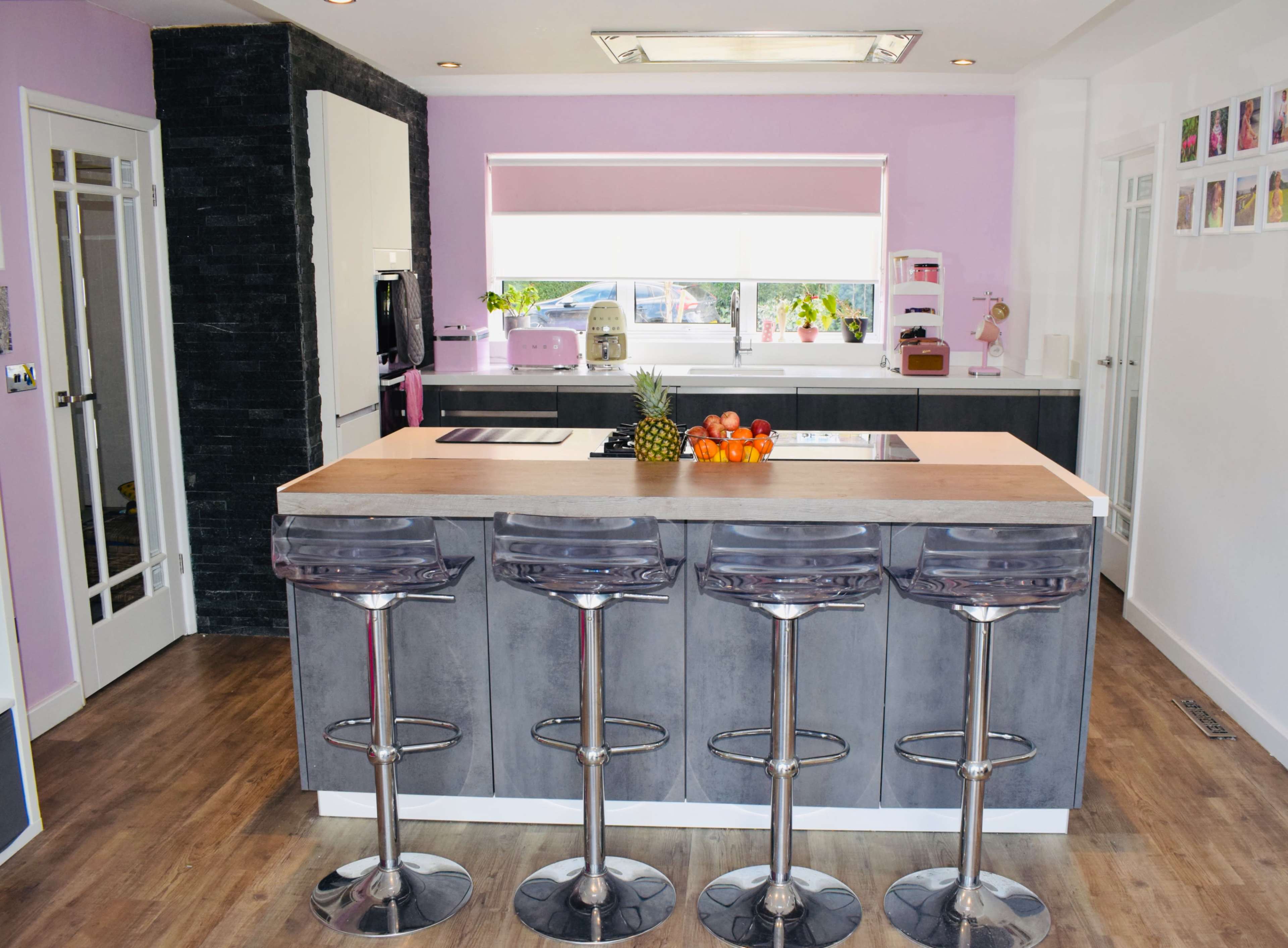 The image features a modern kitchen with a central island, four chrome bar stools, and a combination of pink and gray cabinetry.