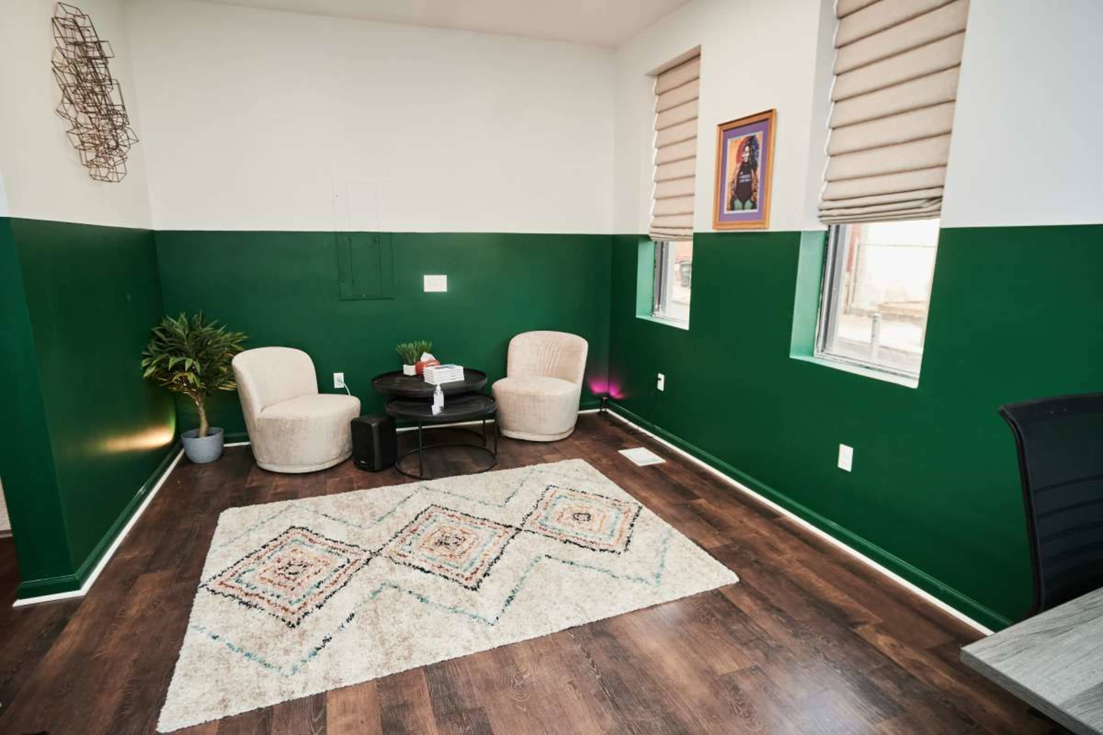 The room features two beige chairs beside a small black table, with green-painted walls and a patterned rug on the wooden floor.
