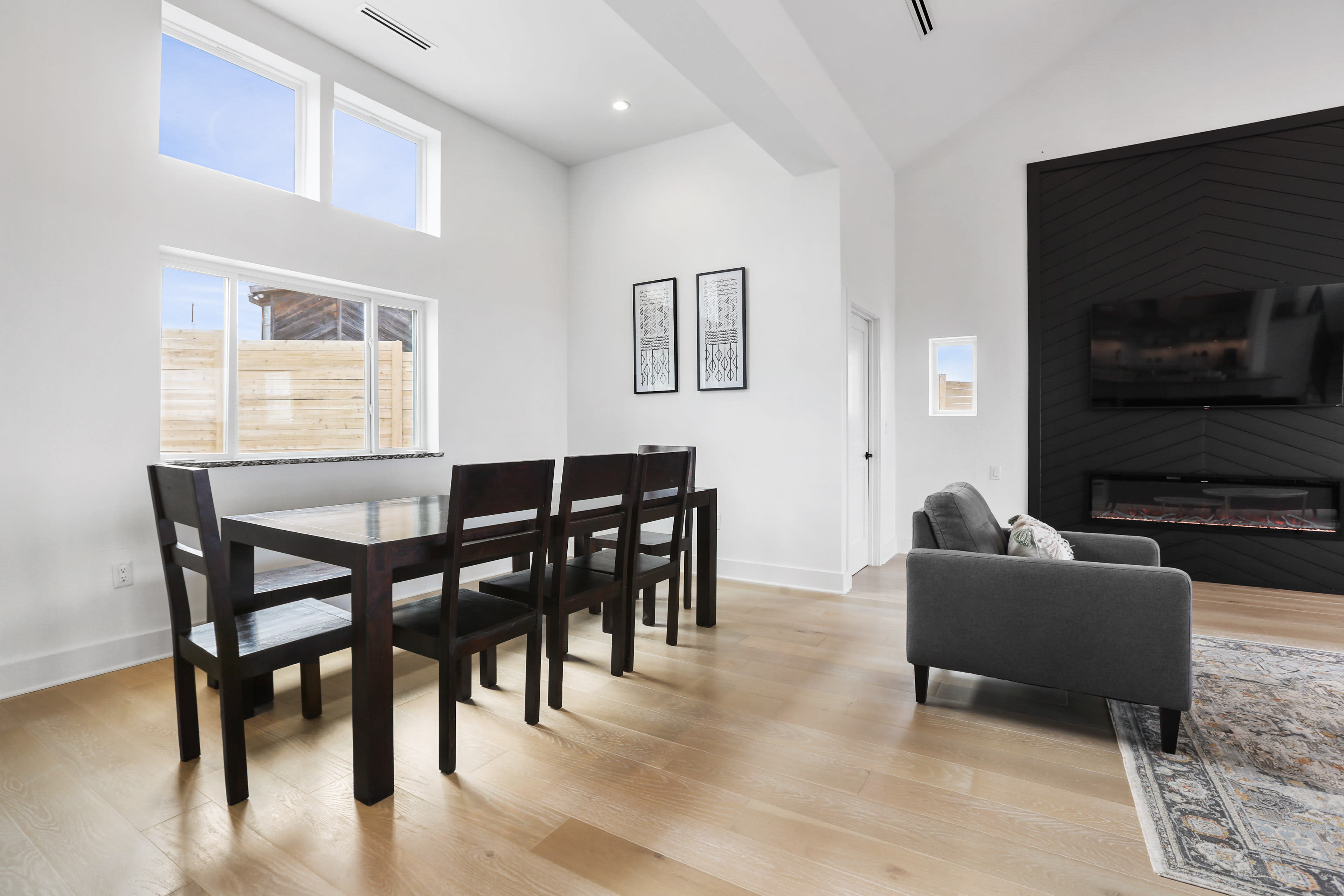 The image shows a modern, light-filled interior featuring a dining table with six chairs beside a gray couch and a wall-mounted television.