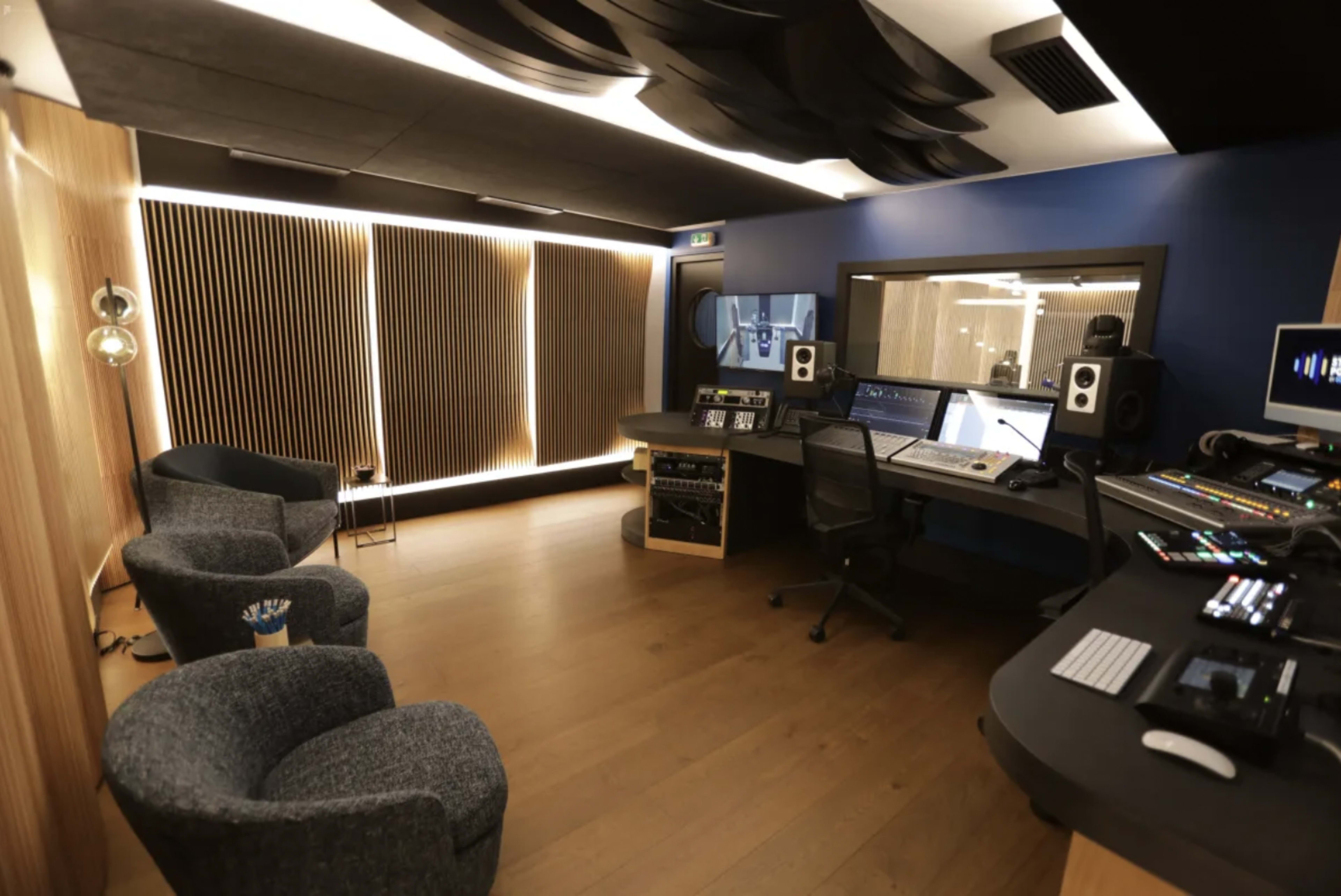 A modern music production studio featuring two armchairs, a large mixing console, speakers, and acoustic paneling along the walls.