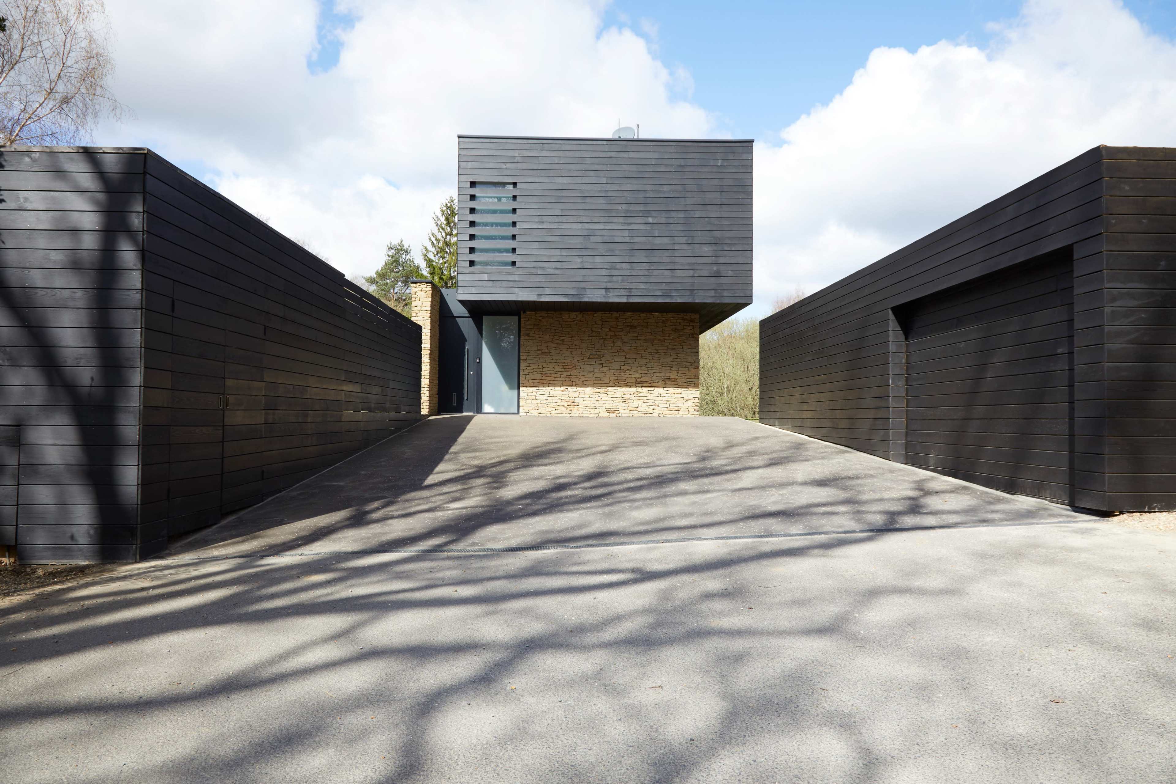 A modern house with a brick ground floor and a dark wooden upper structure, flanked by two wooden fences on either side of a gravel driveway.