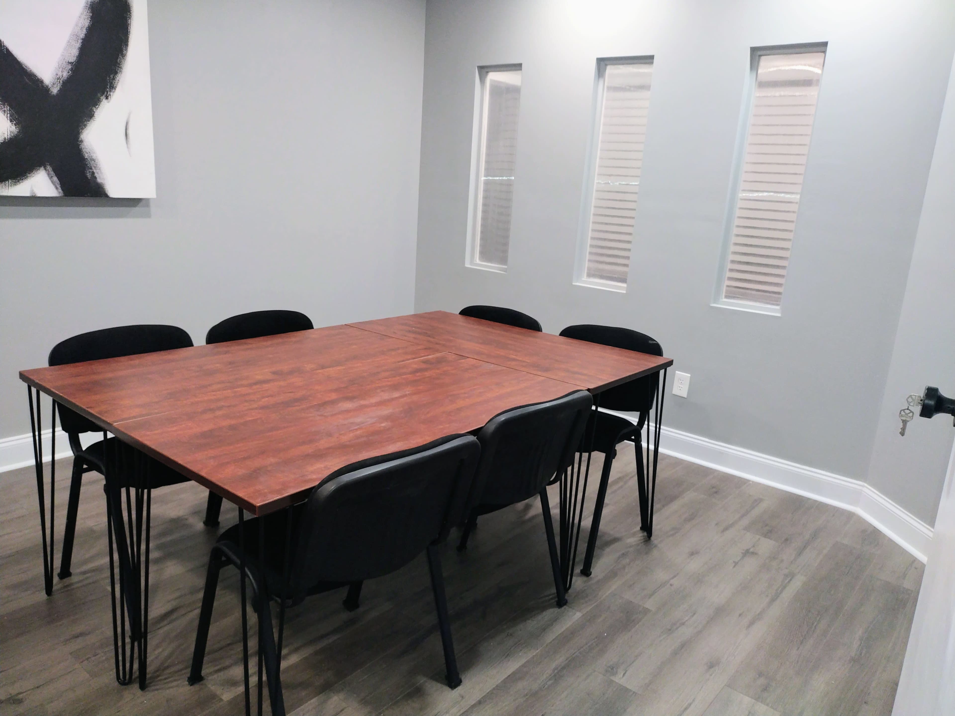 A conference room features a large wooden table surrounded by six black chairs, with three narrow windows on the wall.