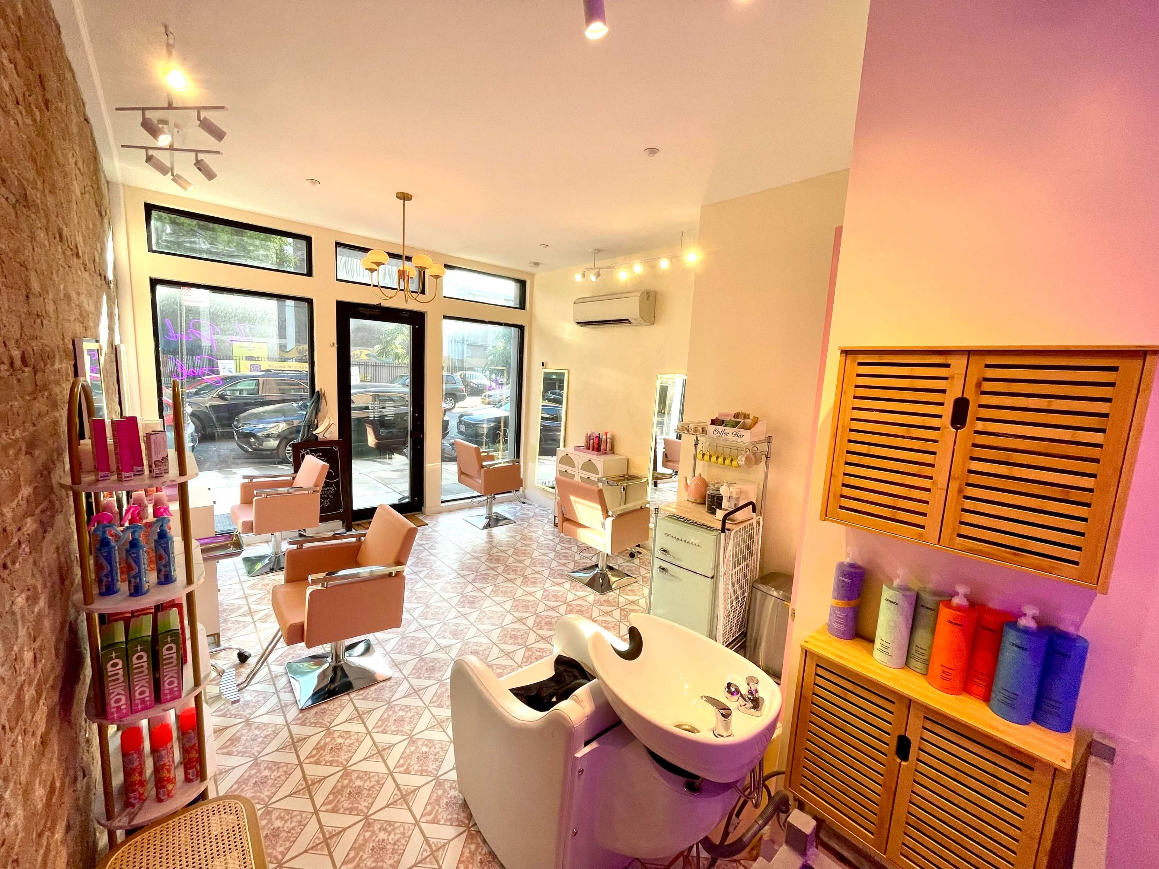 A modern hair salon with several styling chairs, a shampoo station, and shelves stocked with hair products, all framed by large windows allowing natural light to enter.