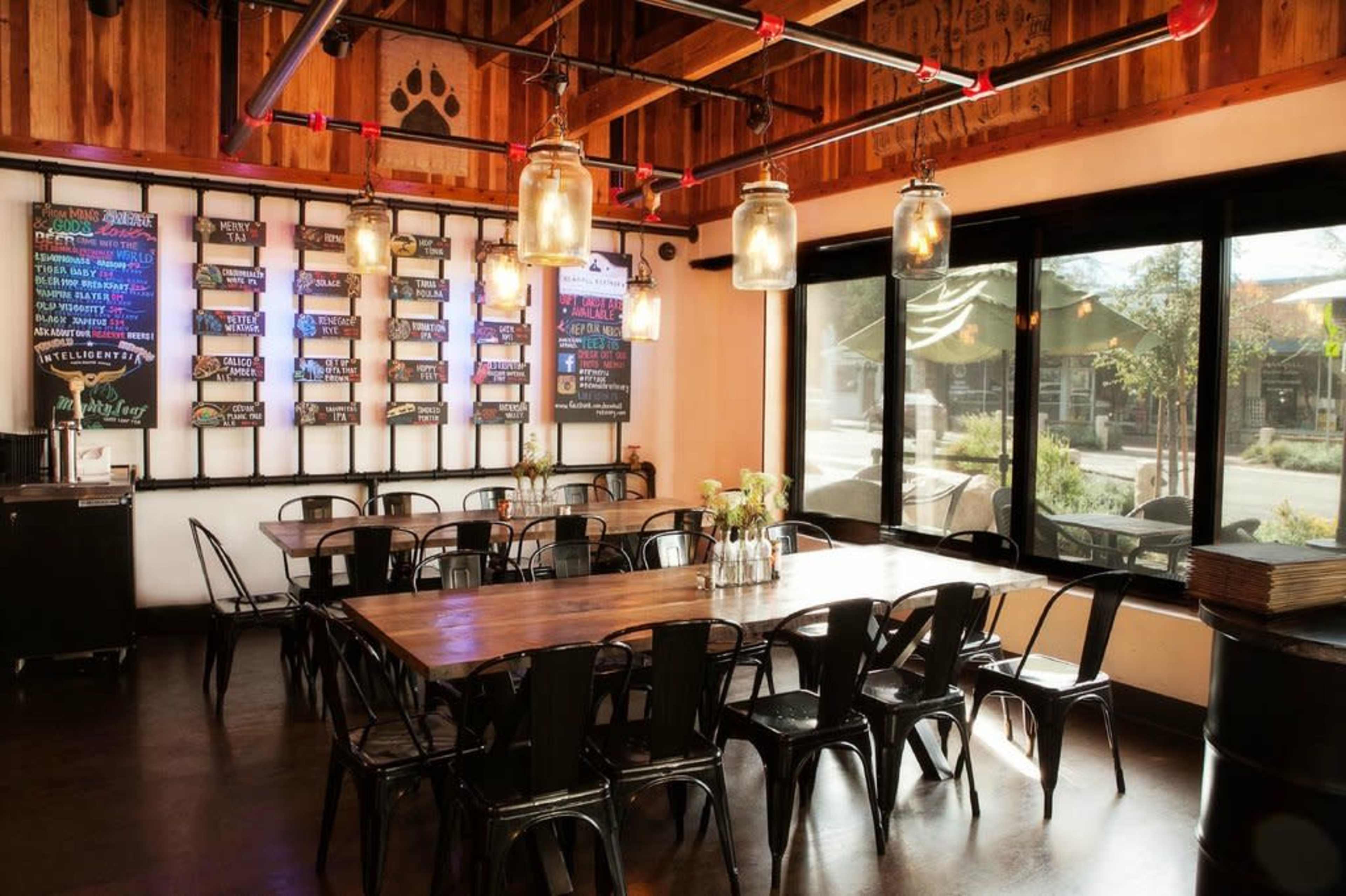 The image shows a spacious restaurant interior with a large wooden table surrounded by black metal chairs, illuminated by hanging glass pendant lights, and featuring a wall with colorful menu boards.