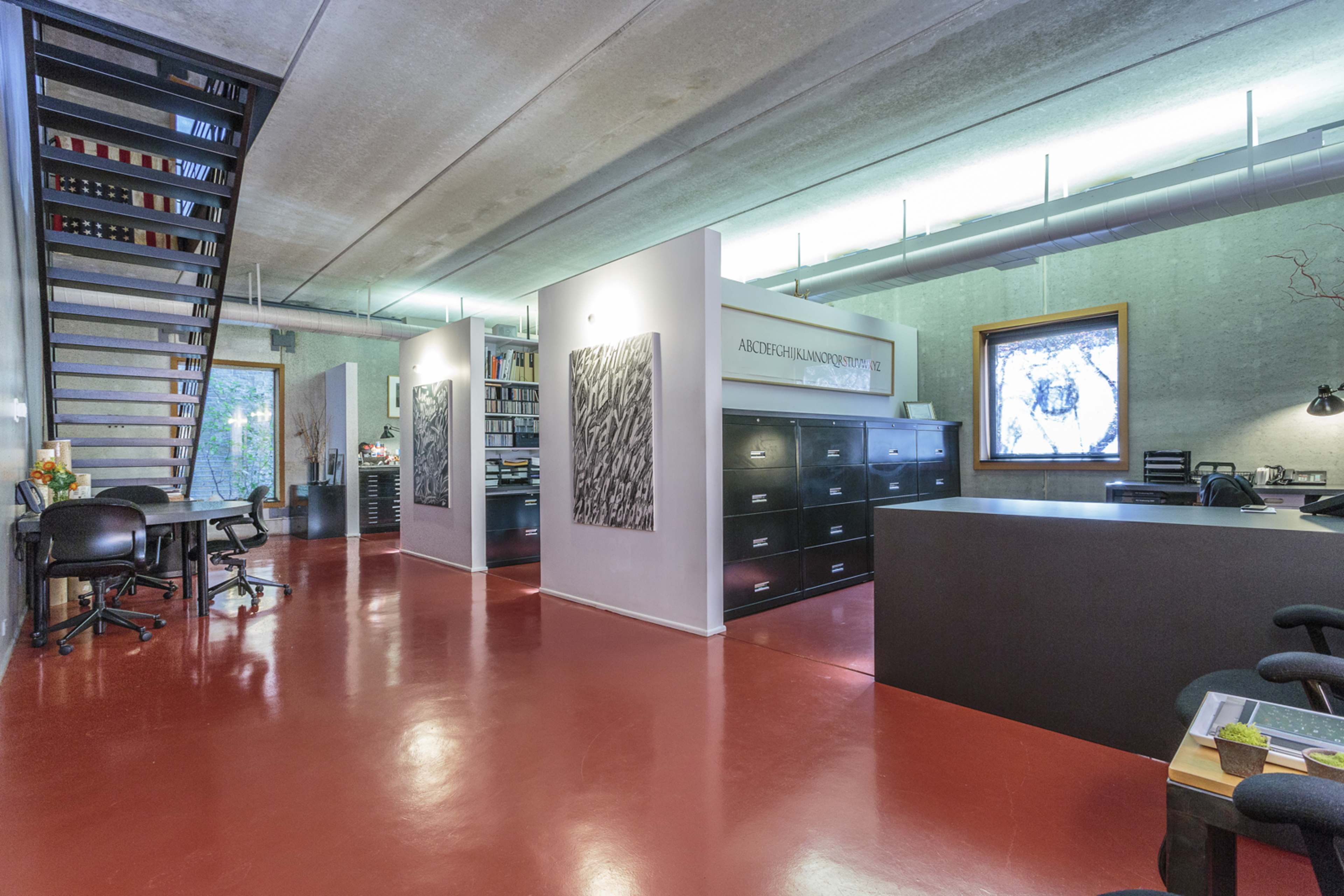 A modern office space with a staircase, filing cabinets, desks, and large windows that allow natural light to enter.