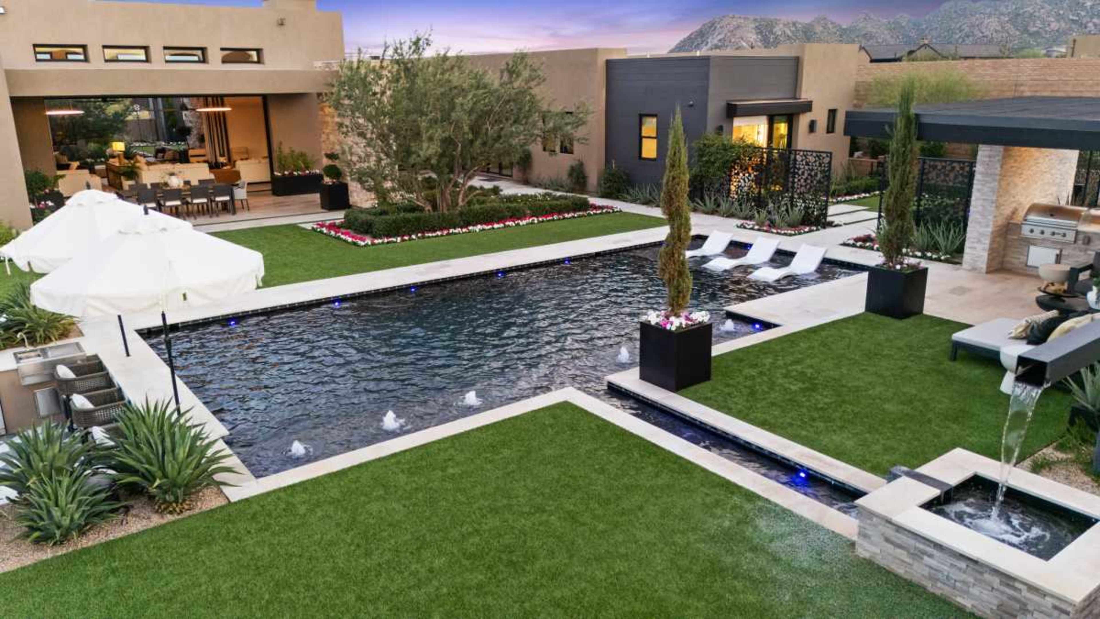 The image features a modern backyard with a swimming pool, lounge chairs, and a dining area surrounded by landscaped greenery and an outdoor kitchen.
