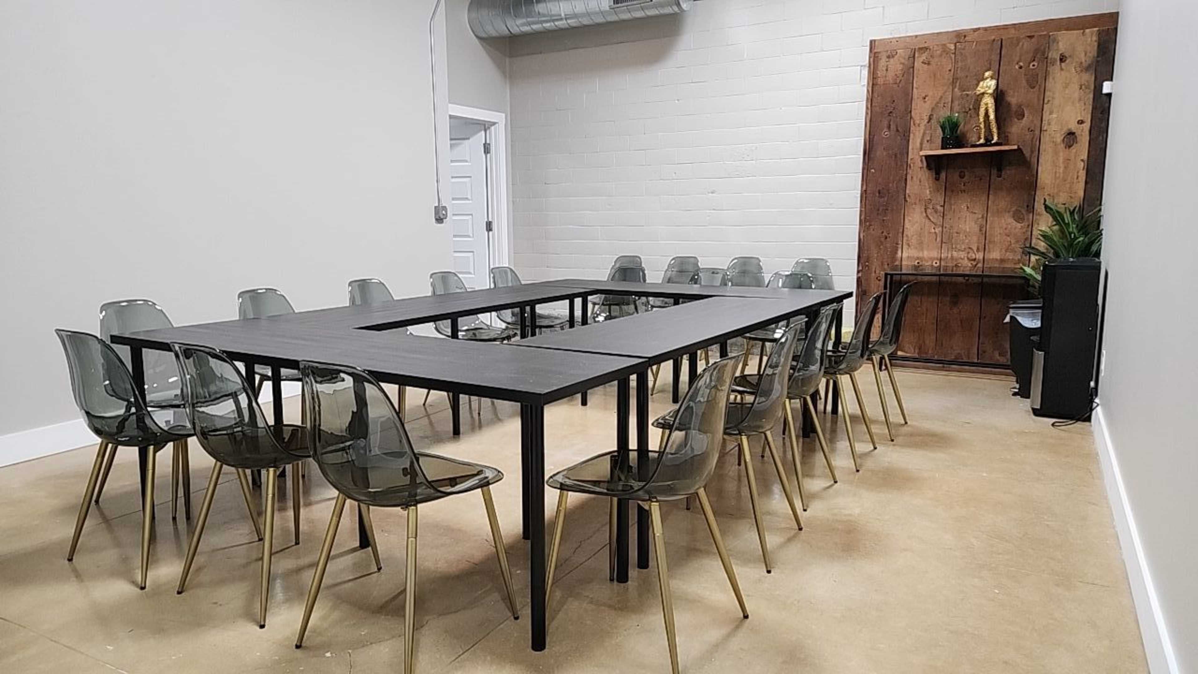 A conference room features a rectangular table surrounded by transparent chairs, with wooden paneling and a plant displayed against the wall.