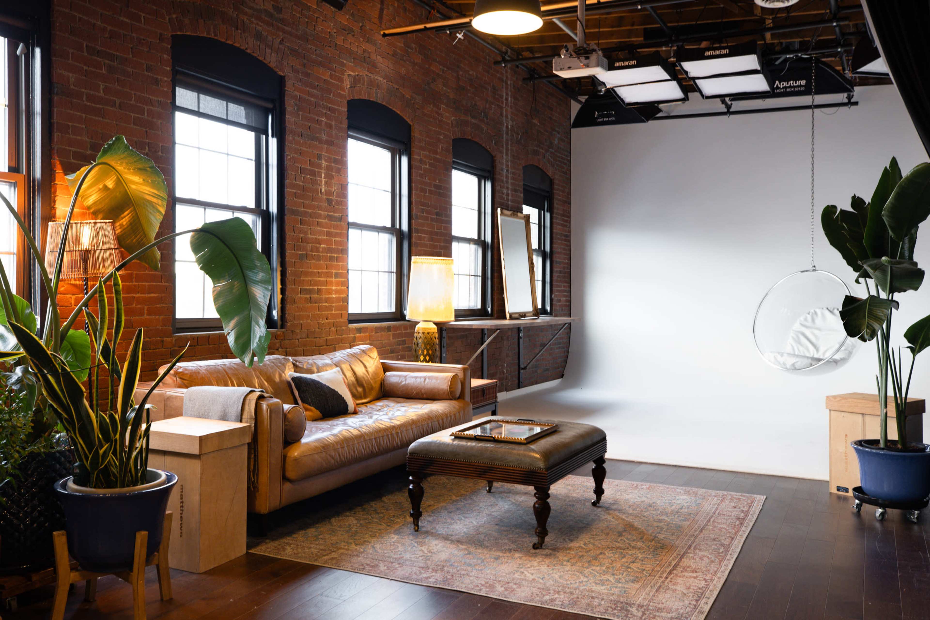The image shows a stylish, well-lit studio with a leather sofa, a decorative rug, potted plants, and a blank backdrop on one side.