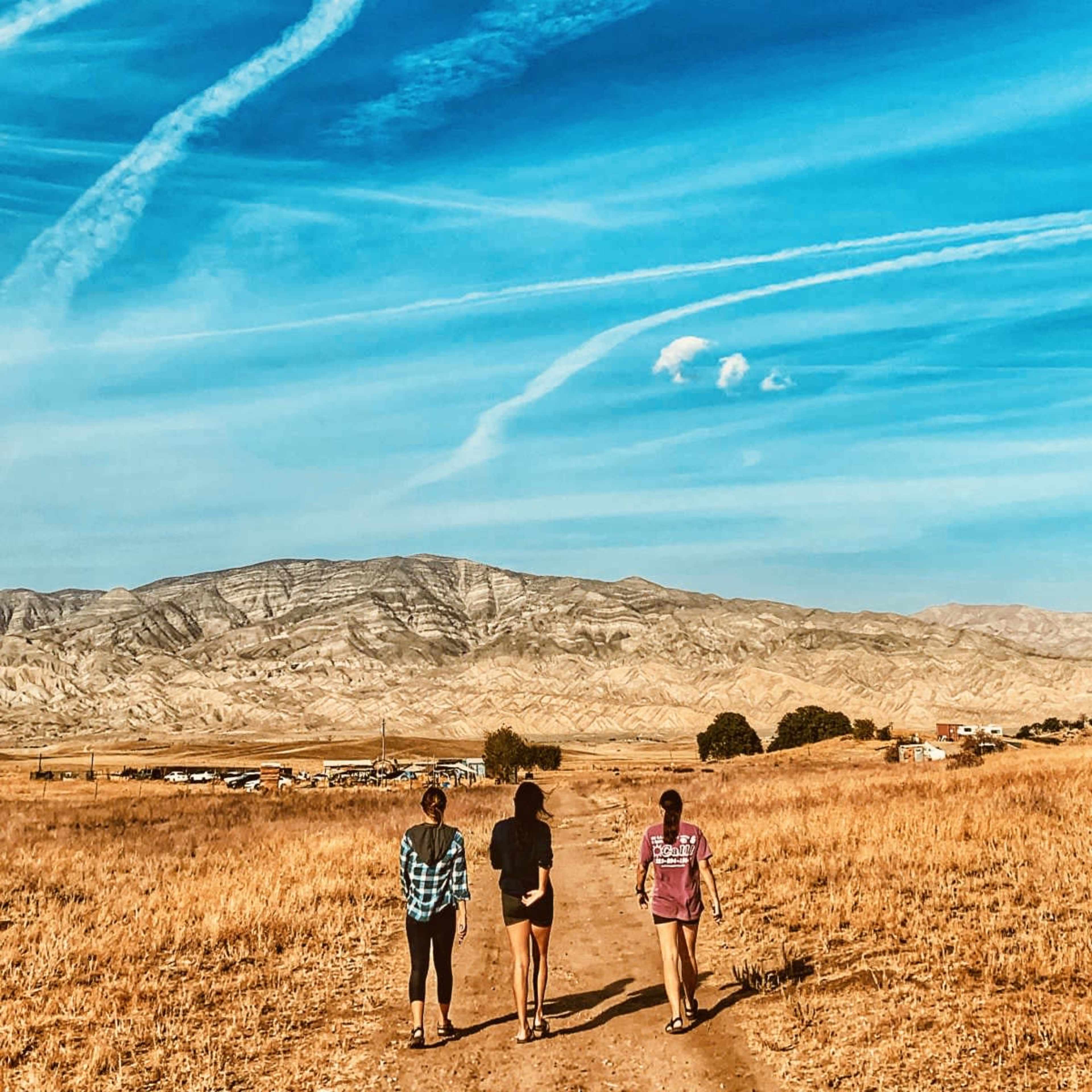 Three individuals walk along a dirt path through a dry, grassy landscape with mountains in the background under a blue sky.