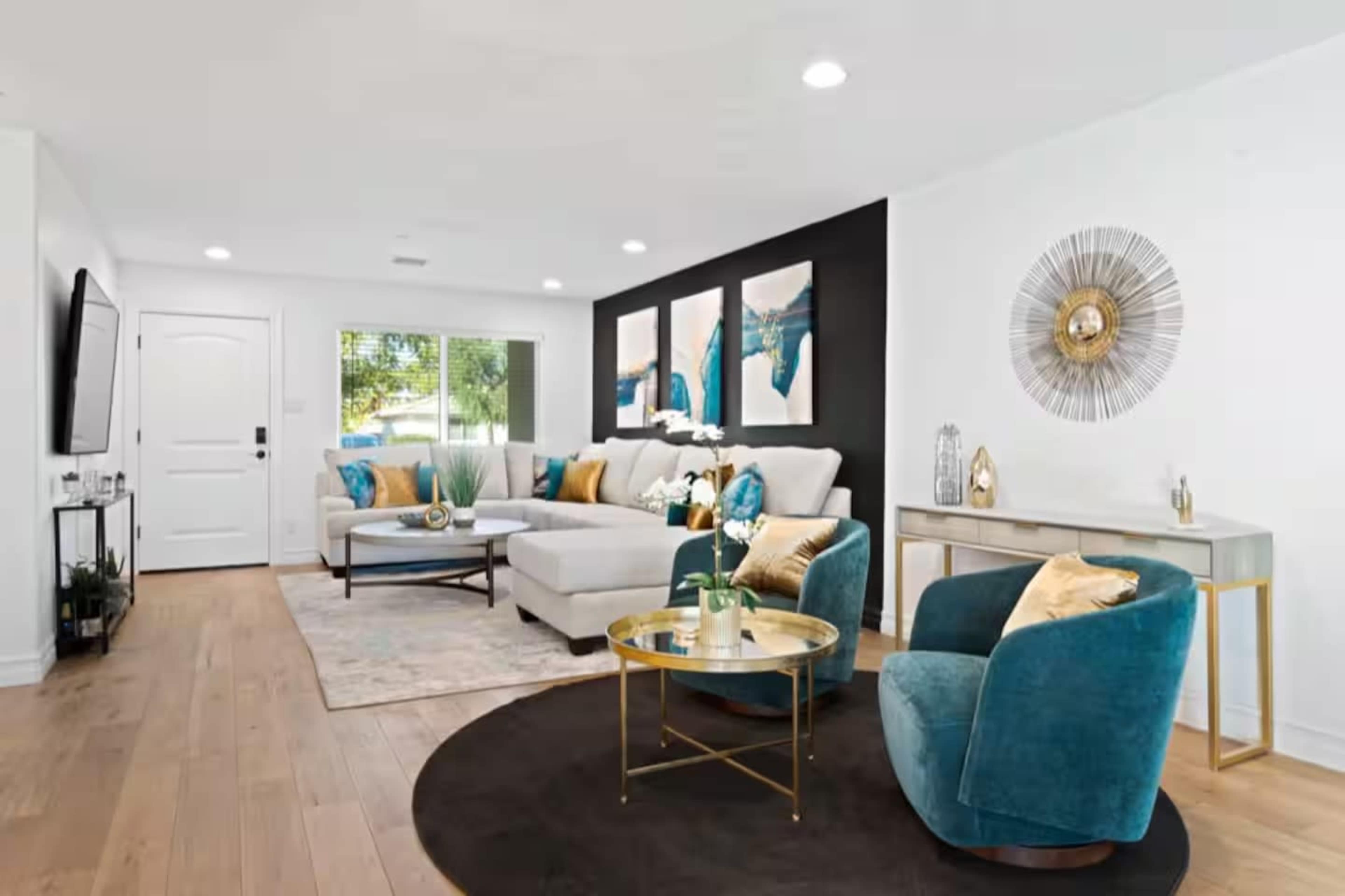 The image shows a modern living room featuring a sectional sofa, two teal armchairs, a round coffee table, and framed artwork on the wall.